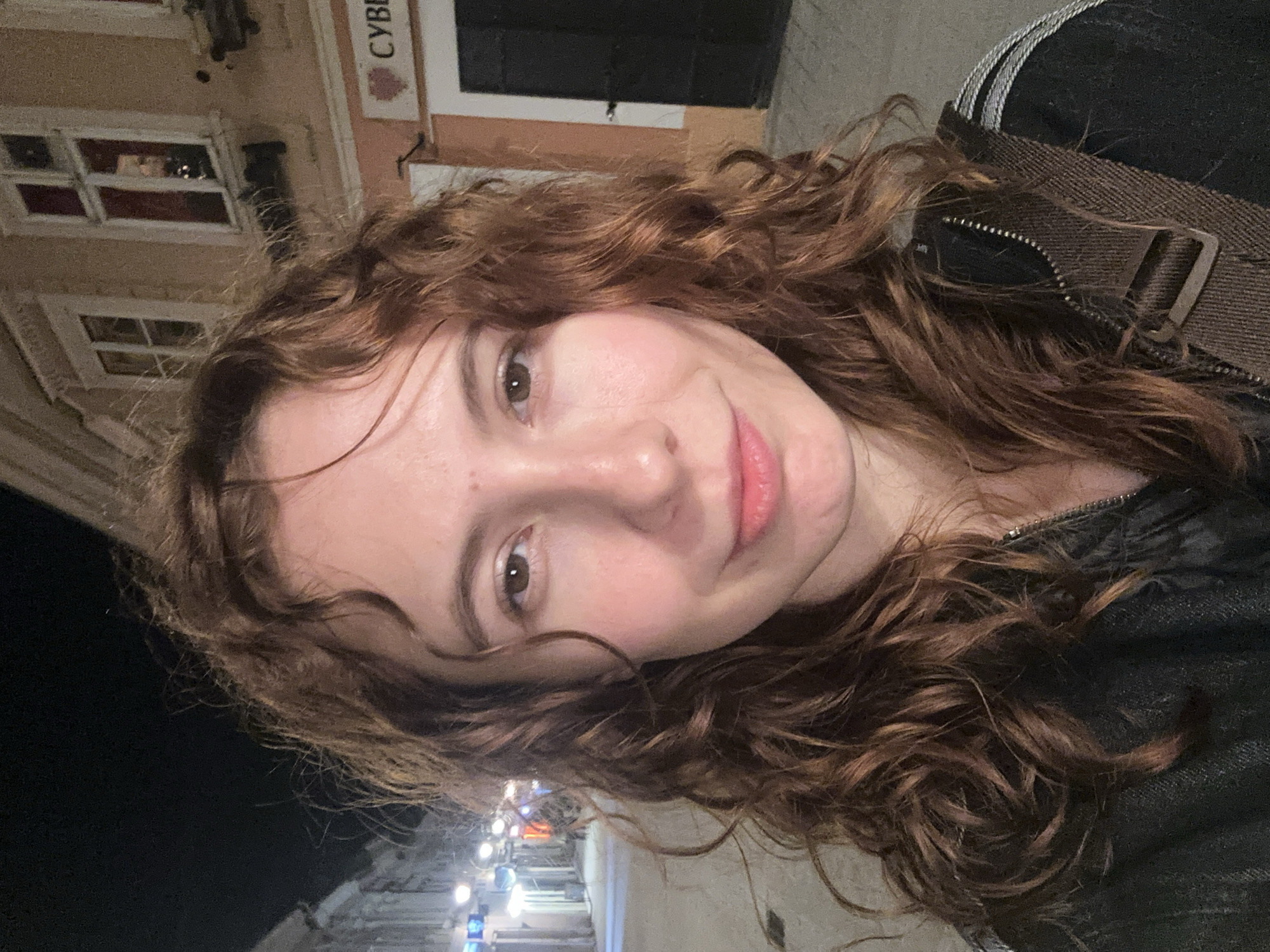 A medium close-up, selfie-style photograph depicts a young woman with light skin and curly brown hair. She is smiling slightly, and her gaze is directed toward the camera. She wears a dark jacket and what appears to be a brown crossbody bag.
The background reveals an urban street scene. Building facades are visible, with windows and architectural details. One building has a sign that reads "CYBE" which could be part of the word "CYBER". Streetlights illuminate the scene.
Based on the architectural style and signage present, the location can be identified as Novi Sad, Serbia.
