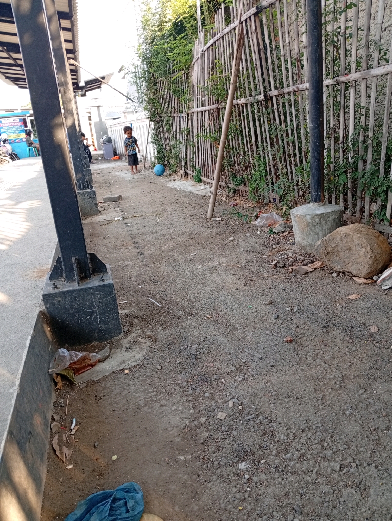 The image depicts an outdoor scene in what appears to be a public space with a dirt and gravel ground. In the mid-ground, a young child, likely a boy, is standing near a bamboo fence, holding a stick and looking towards a blue ball on the ground. To the left, there is a covered structure with black pillars and a metal roof, suggesting it might be a shelter or part of a building. A few people are visible in the background near a blue sign and some motorcycles, indicating a potential transportation hub or waiting area. The environment is characterized by natural vegetation growing along the fence, scattered debris on the ground including leaves and plastic bags, and a large rock near a concrete base. The overall impression is a somewhat unkempt outdoor area. Without further context, pinpointing the exact location within Karawang, Indonesia, is not possible.