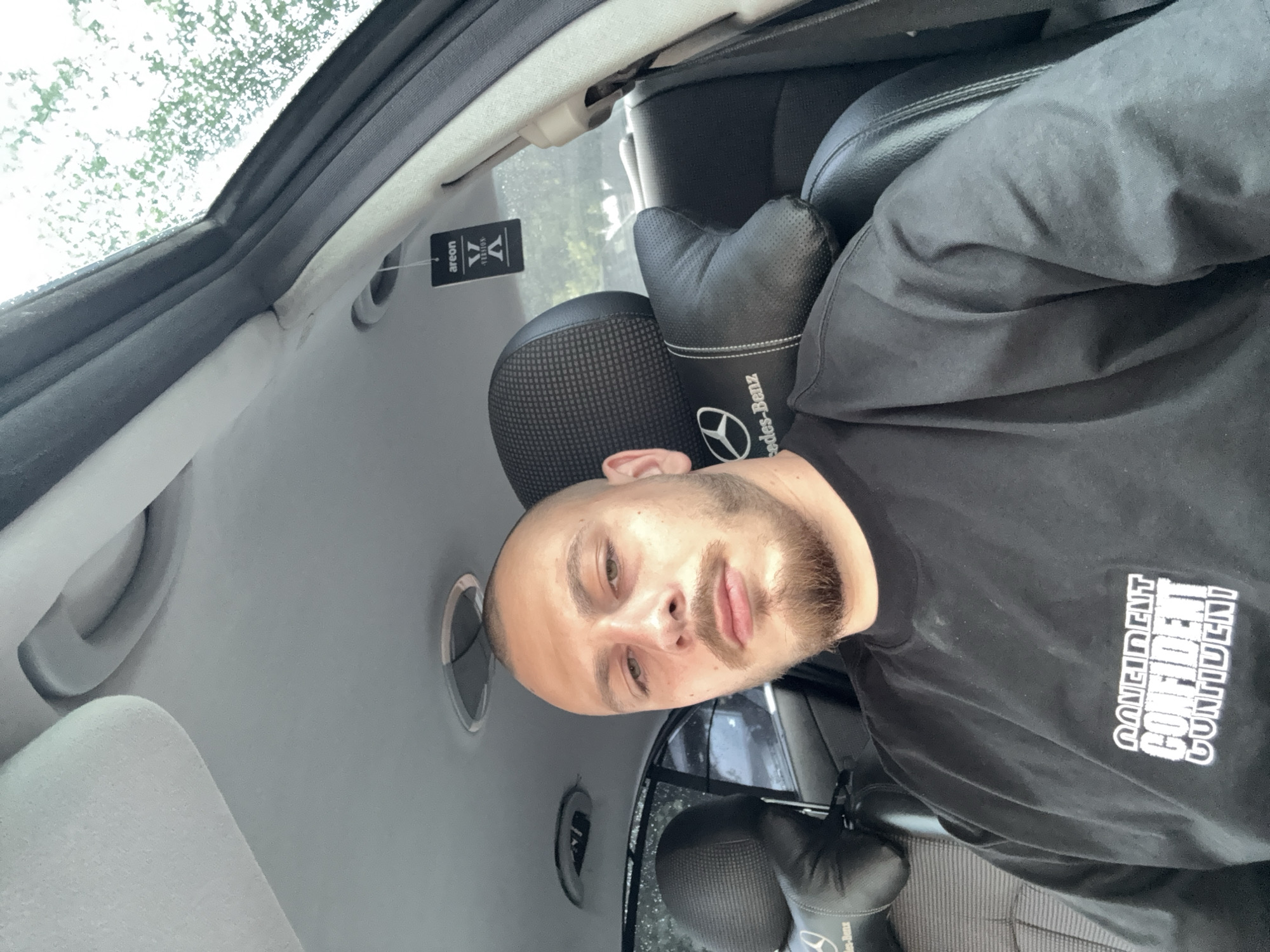A male individual is depicted in a self-portrait, seated within the interior of a vehicle. The subject has a shaved head, a light brown beard, and a neutral facial expression. He wears a black crew-neck t-shirt adorned with a white graphic on the chest that features the word "CONFIDENT" stacked multiple times in a stylized font. The vehicle's interior exhibits black leather seats with patterned fabric inserts and a light gray headliner. A black leather neck pillow, embroidered with the "Mercedes-Benz" logo, is attached to the seat. A black, rectangular "areon V VERSION" air freshener hangs from the overhead console area near the right A-pillar. A partially open sunroof is visible above, revealing blurred green foliage and a bright sky with evident moisture or raindrops on the glass. The scene is located in the city of Stara Zagora, Bulgaria.
