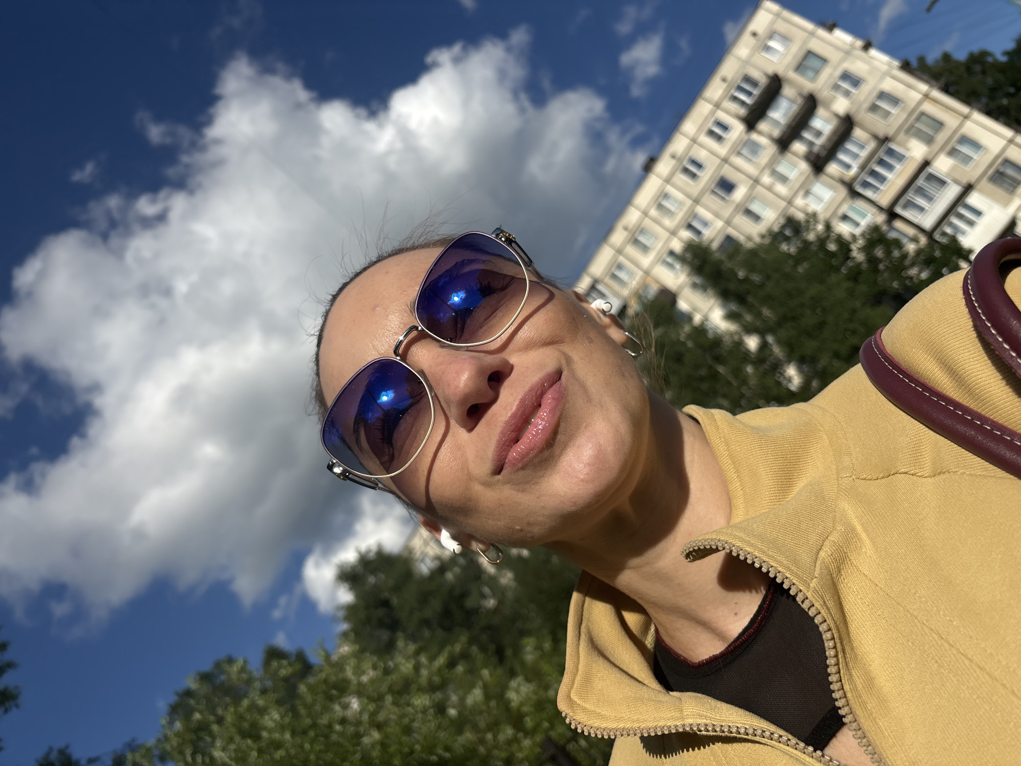 In this close-up, low-angle selfie, a woman looks up at the camera, her face framed by sunglasses that reflect the bright blue sky and fluffy white clouds. Her lips are slightly parted, and she appears to be smiling faintly. She is wearing a yellow zip-up jacket over a dark top. A small burgundy strap, likely from a bag, is visible over her shoulder. In the background, a tall, beige apartment building with many windows is tilted diagonally in the frame, suggesting an unusual perspective or a slight tilt of the camera. Green trees are visible in the lower part of the image, and the overall lighting indicates it is daytime, likely sunny with some cloud cover. The overall impression is a casual, outdoor snapshot.