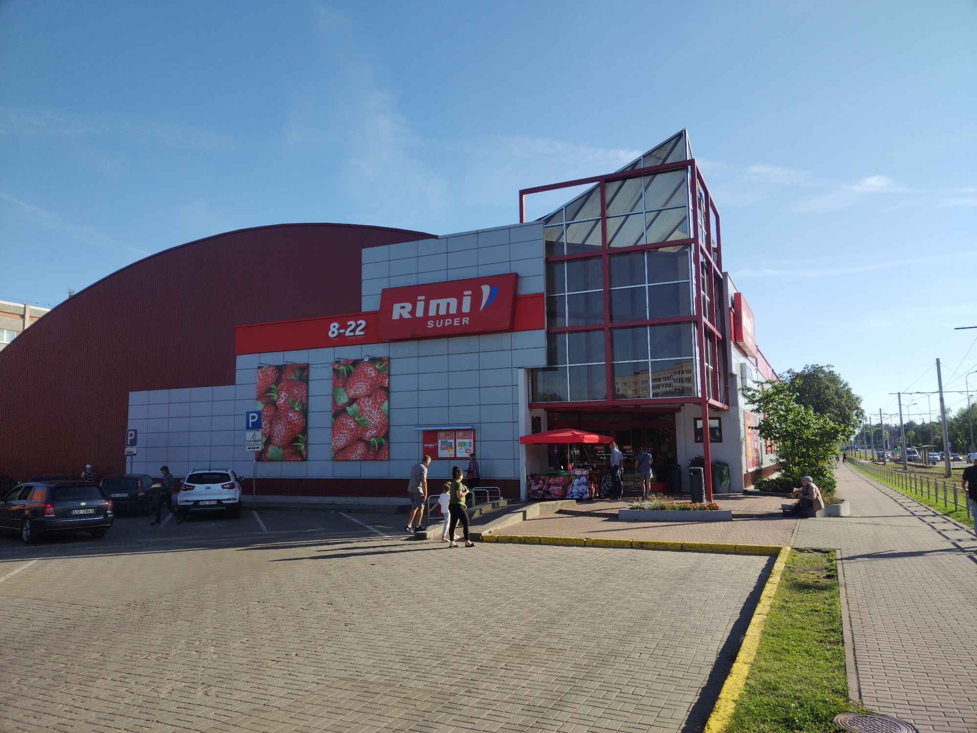 The image shows a Rimi supermarket in Daugavpils, Latvia. The building is modern, with a large red curved roof and a glass-fronted section. The Rimi logo is prominently displayed above the entrance, along with the operating hours "8-22". Large images of strawberries are visible on the side of the building.
In the foreground, a paved parking lot is visible with several cars parked. A few people are walking in the parking lot and on the sidewalk. Some people are sitting on benches outside the store. In the background, a street with traffic and power lines can be seen. The sky is blue and clear. The overall scene depicts a typical day at a supermarket in a city.