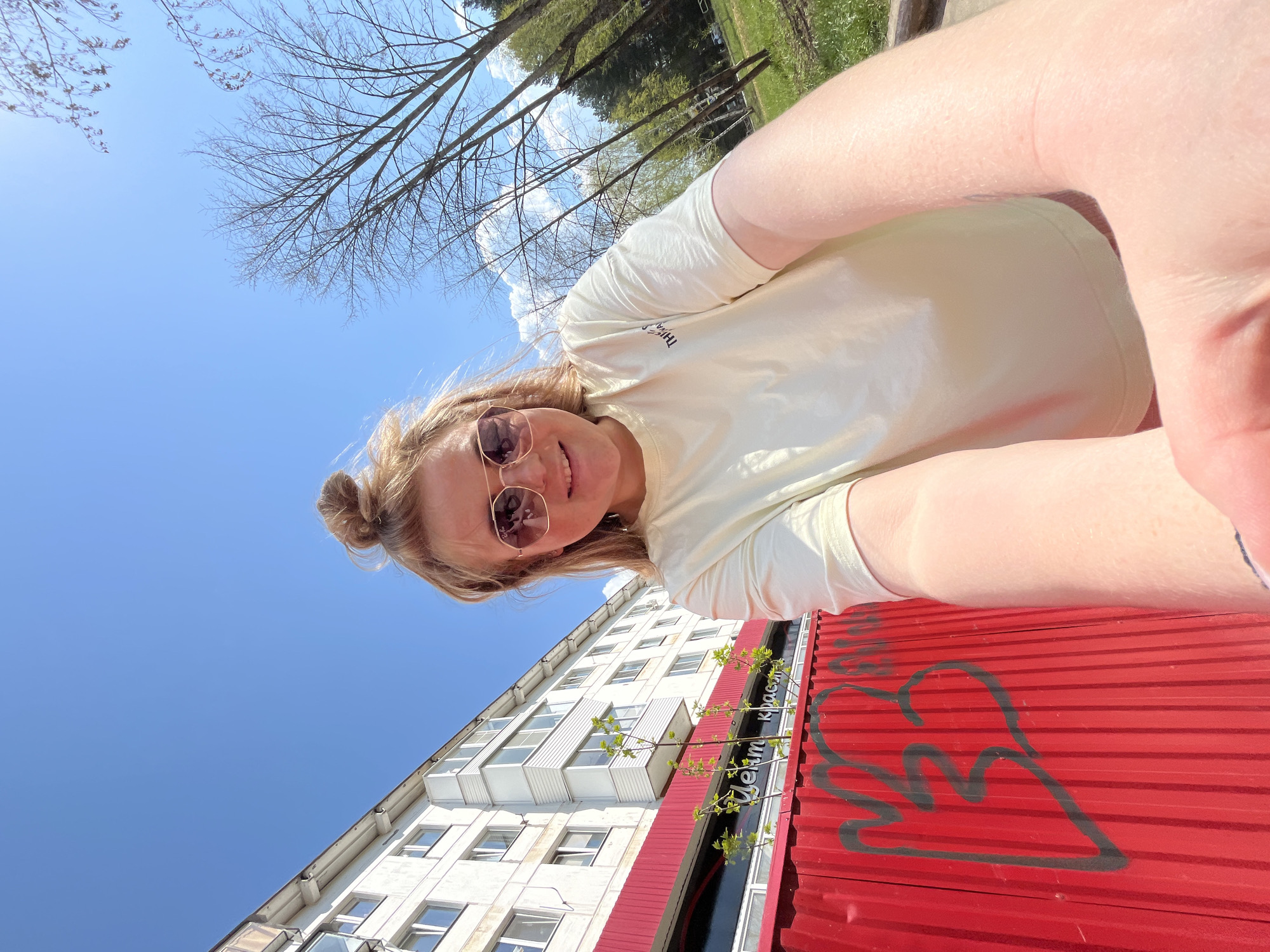 The image depicts a selfie taken by a woman with blonde hair pulled into a bun, wearing sunglasses and a light-colored t-shirt. The background shows a building with white walls and multiple windows, as well as a red corrugated metal fence or wall with graffiti. Above the building and the woman are blue skies and some bare tree branches. The photograph was taken on a sunny day in the city of Chaikovsky, Russia.