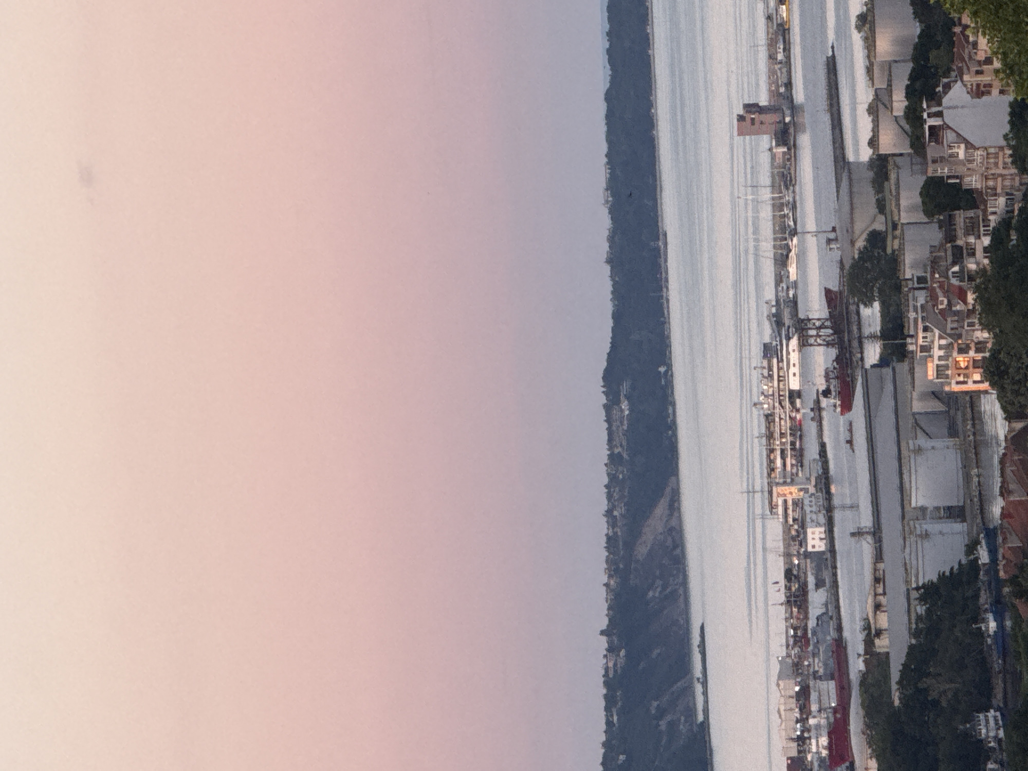 The image depicts a panoramic view of a harbor and a coastal town at dusk. The sky is a gradient of soft pink and lavender, transitioning to a pale blue towards the horizon. In the foreground, a cluster of white industrial storage tanks are visible, along with some buildings with red-tiled roofs. Beyond these, the harbor is filled with various vessels, including large cargo ships, smaller boats, and sailboats with tall masts. A prominent red cargo ship is moored in the middle of the frame. Further in the distance, a town is nestled along a lush green hillside that slopes down to the sea. The water in the harbor and sea is calm and reflects the ambient light. A distinctive reddish-brown, multi-story structure with a tower-like top stands out in the harbor area. The overall impression is one of a bustling port city at the quiet close of day.