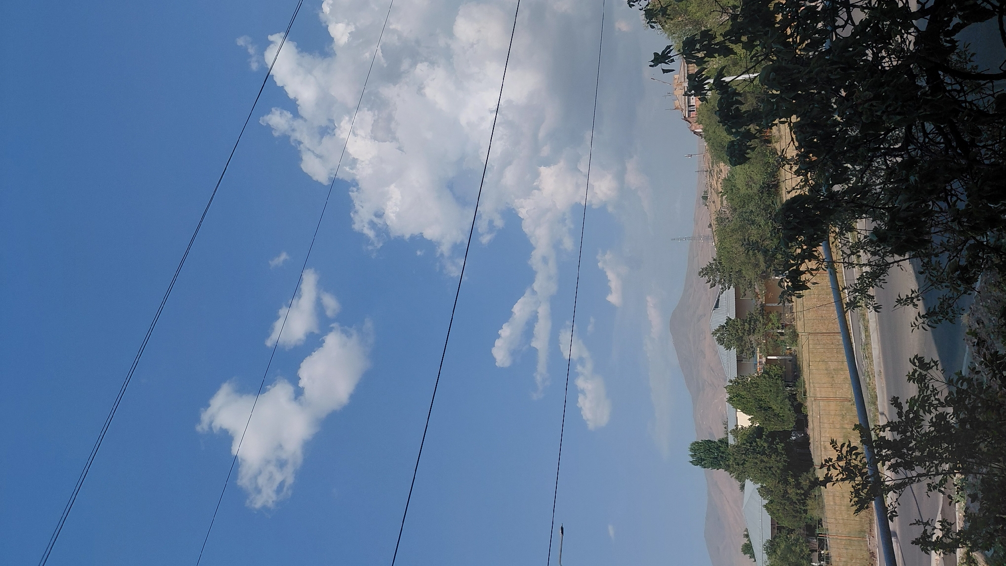 The image depicts a clear, bright sky with scattered white clouds. Three parallel black wires stretch across the upper portion of the frame. Below the sky, a hazy landscape unfolds, featuring rolling hills in the distance under a muted blue sky. In the mid-ground, there are several buildings with grey roofs and beige walls, surrounded by lush green trees. A tall, slender tower is visible on the right side of the mid-ground. In the foreground, a dark, leafy tree obscures part of the view. A metal fence runs horizontally across the middle of the image, separating the buildings from a road below. The road appears to be asphalt and is empty of vehicles. The scene suggests a suburban or semi-rural environment.