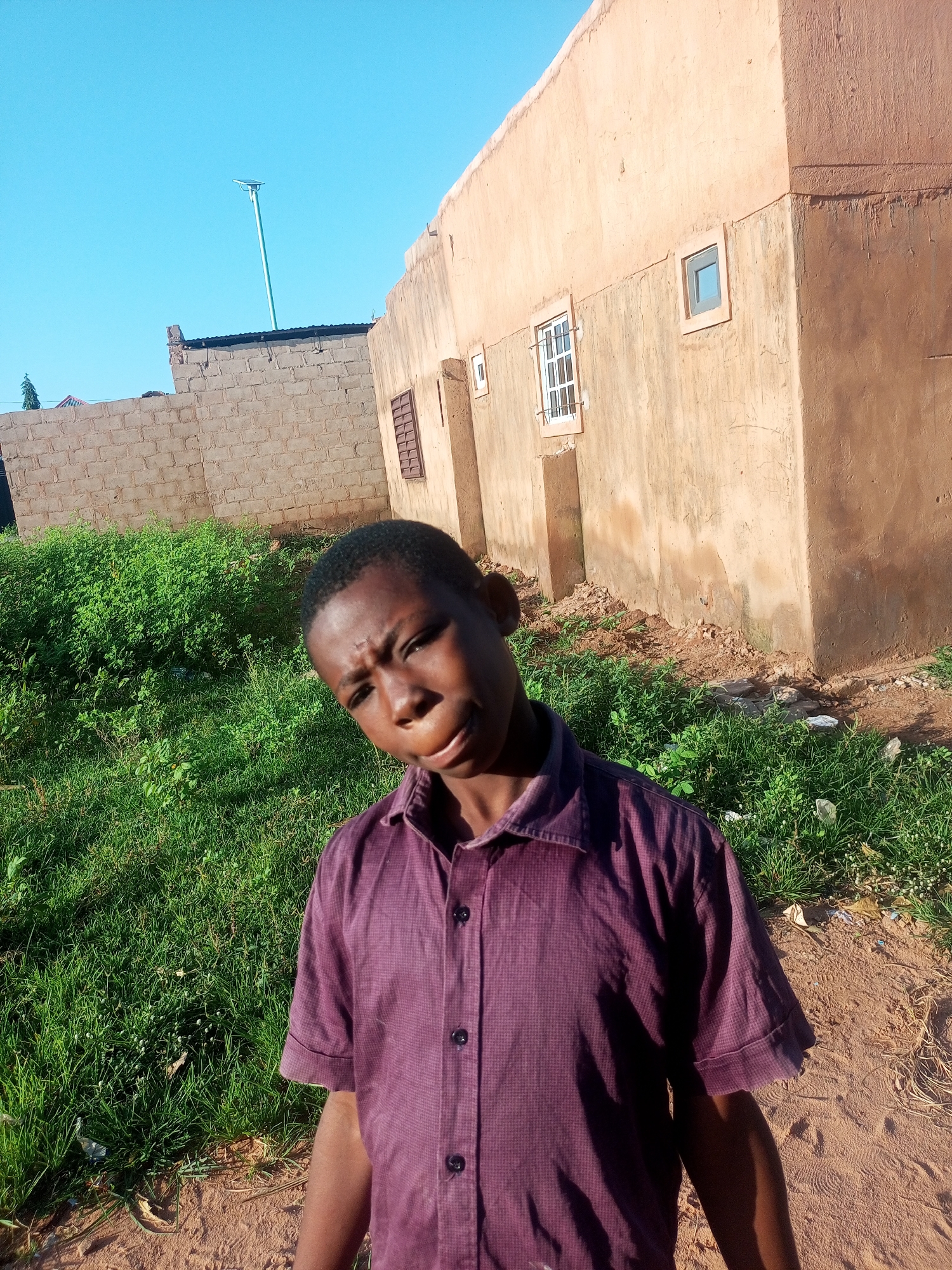 In the city of Tempure, Nigeria, a young boy with dark skin and short black hair stands in front of a partially constructed building. He is wearing a short-sleeved, collared purple shirt. The building is made of beige stucco and features two windows, one with a white frame and bars, and another smaller, darker window. To the left of the building is a wall constructed of light-colored bricks, and beyond that, a patch of vibrant green grass. The ground in front of the boy is a reddish-brown dirt path. The sky above is a clear, bright blue.