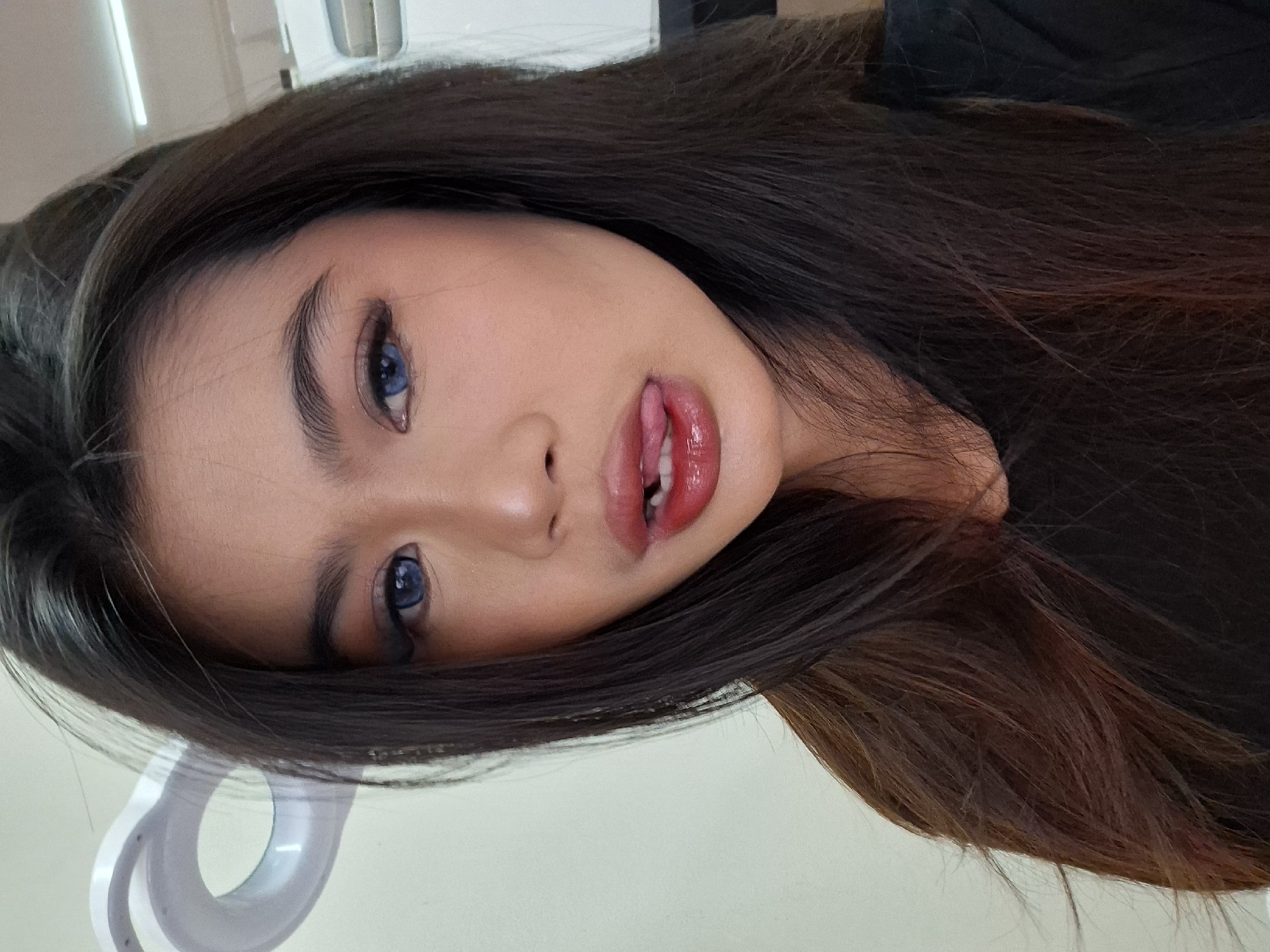 A young woman with long, dark brown hair and bright blue contact lenses is looking directly at the camera. She has on bold makeup, including winged eyeliner, eyeshadow, and bright red lipstick. Her tongue is sticking out slightly, touching her lower lip. She is wearing a black top. In the background, a ring light is visible, suggesting the photo was taken indoors, possibly for a selfie or to showcase makeup. The setting appears to be a room with plain, light-colored walls and overhead lighting. The image is a close-up, focusing on the woman's face and upper body. There is no discernible information about the location being Kuta, Indonesia.