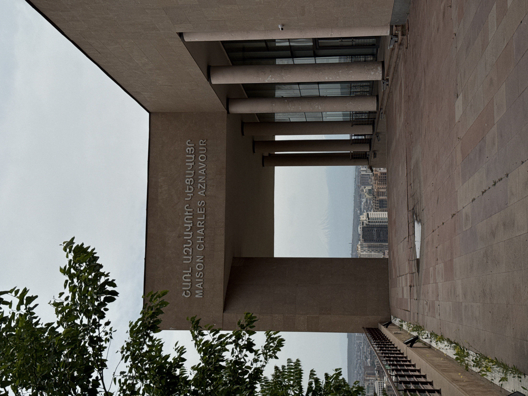 The image shows the Maison Charles Aznavour in Yerevan, Armenia. The building features a minimalist architectural design with a prominent concrete structure bearing the name of Charles Aznavour in both Armenian and English. A series of cylindrical columns support an overhanging structure, framing a view of the city skyline and a distant mountain. The ground is paved with stone tiles, and greenery is visible in the foreground. The sky is overcast.