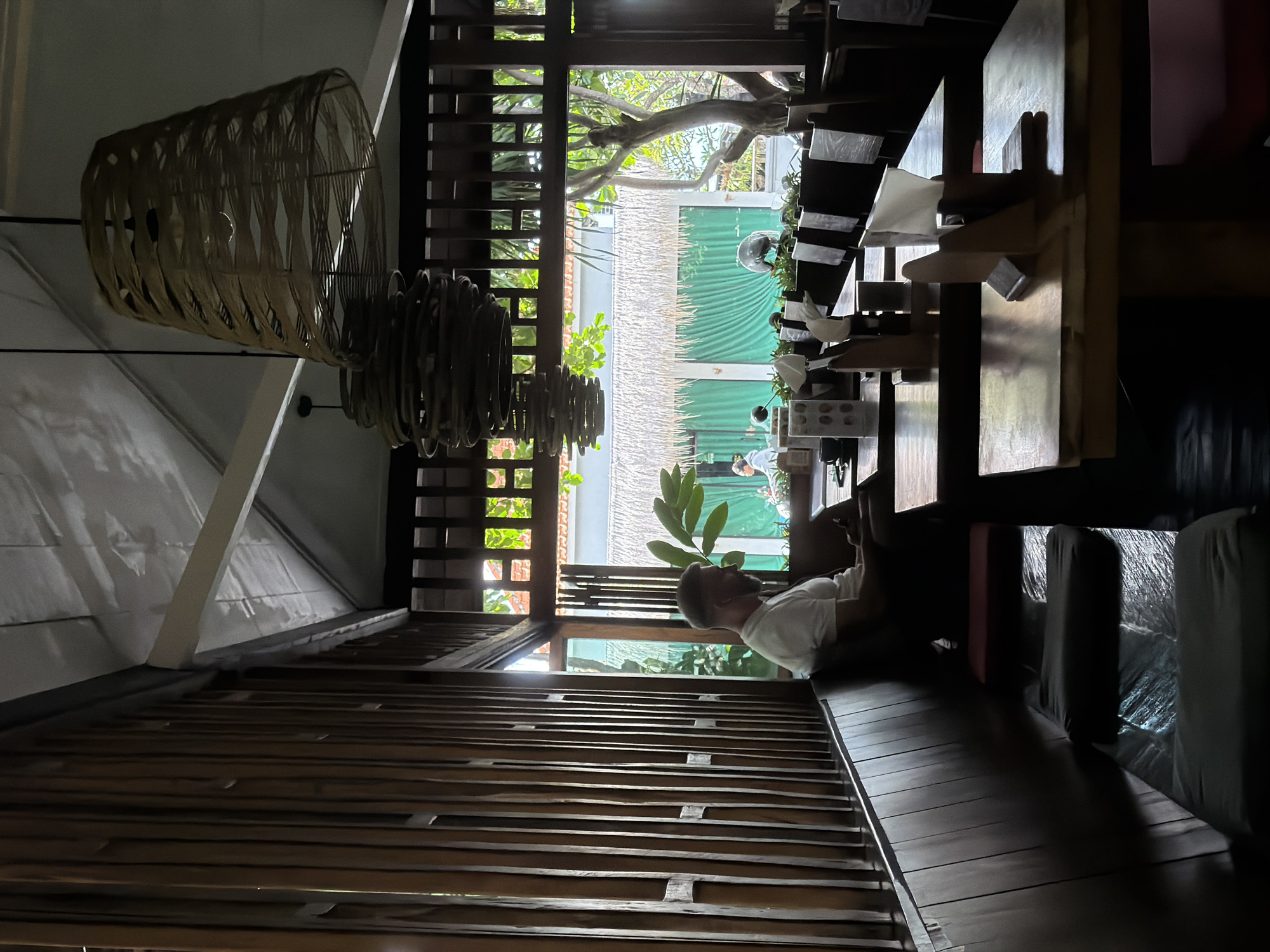 The image depicts an indoor scene, possibly a café or lounge in North Kuta, Indonesia, as suggested by the architectural style and vegetation visible through the window. The foreground features a wooden slat wall and ceiling with a large, woven pendant light fixture hanging from the ceiling.
In the background, a man is sitting on a cushioned bench, wearing a white t-shirt. He is looking down at an object in his hands, possibly a phone. The area behind him is well-lit, with an outdoor view framed by wooden lattice windows. This view includes tropical foliage and a glimpse of the building exterior, possibly a restaurant's or shop's decor. Tables and chairs are arranged in the distance, suggesting a dining or gathering space.
