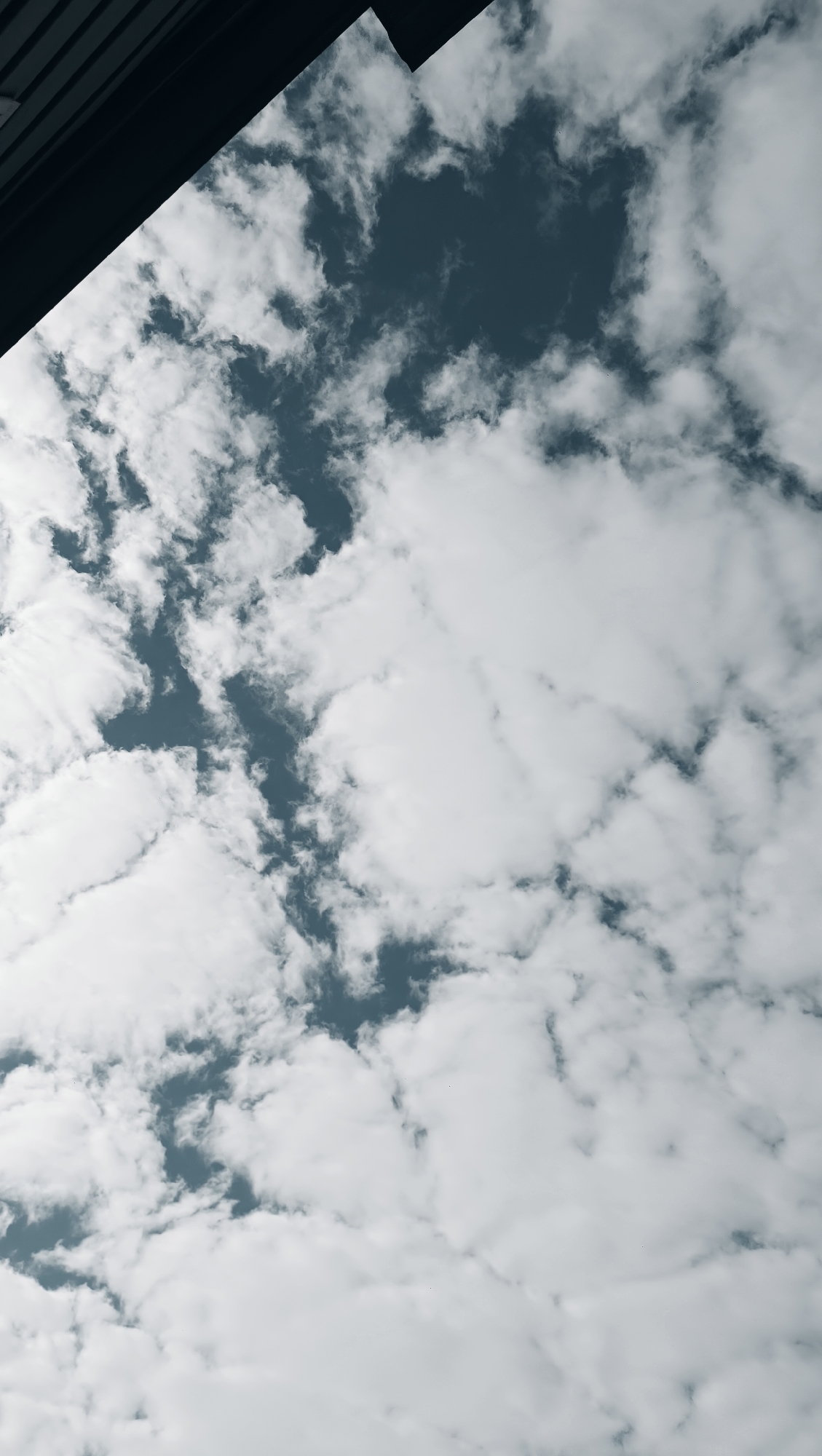 The image shows a dark, angled architectural element in the upper left corner, contrasting with a vast expanse of cloudy sky. The clouds are white and fluffy, interspersed with dark blue patches of clear sky. The perspective is from below, looking upwards. The scene is a sky view, with no people, animals, or specific objects identifiable as being in the city of Karsha, Russia. The image is a photograph of a cloudy sky with a portion of a dark building or structure at the top left.