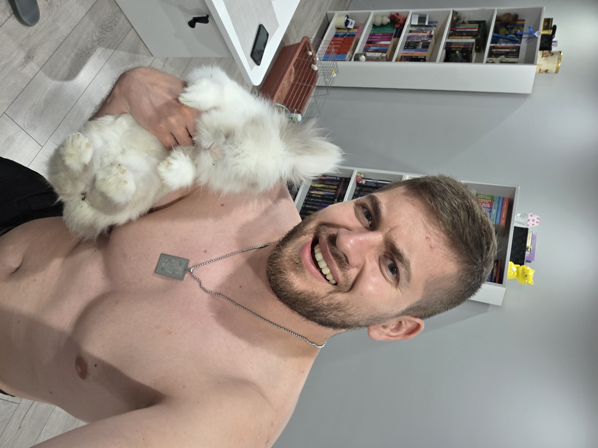 A fair-skinned man with a beard and short brown hair is holding a white rabbit. The man is topless and wearing a silver necklace with a rectangular pendant. He is smiling and looking at the camera. The rabbit is held upside down. Behind the man is a wall with shelves filled with books, movies, and other objects. In the background is a white table with a brown rabbit cage. There is no verifiable evidence of the location being Yagodovo, Bulgaria.