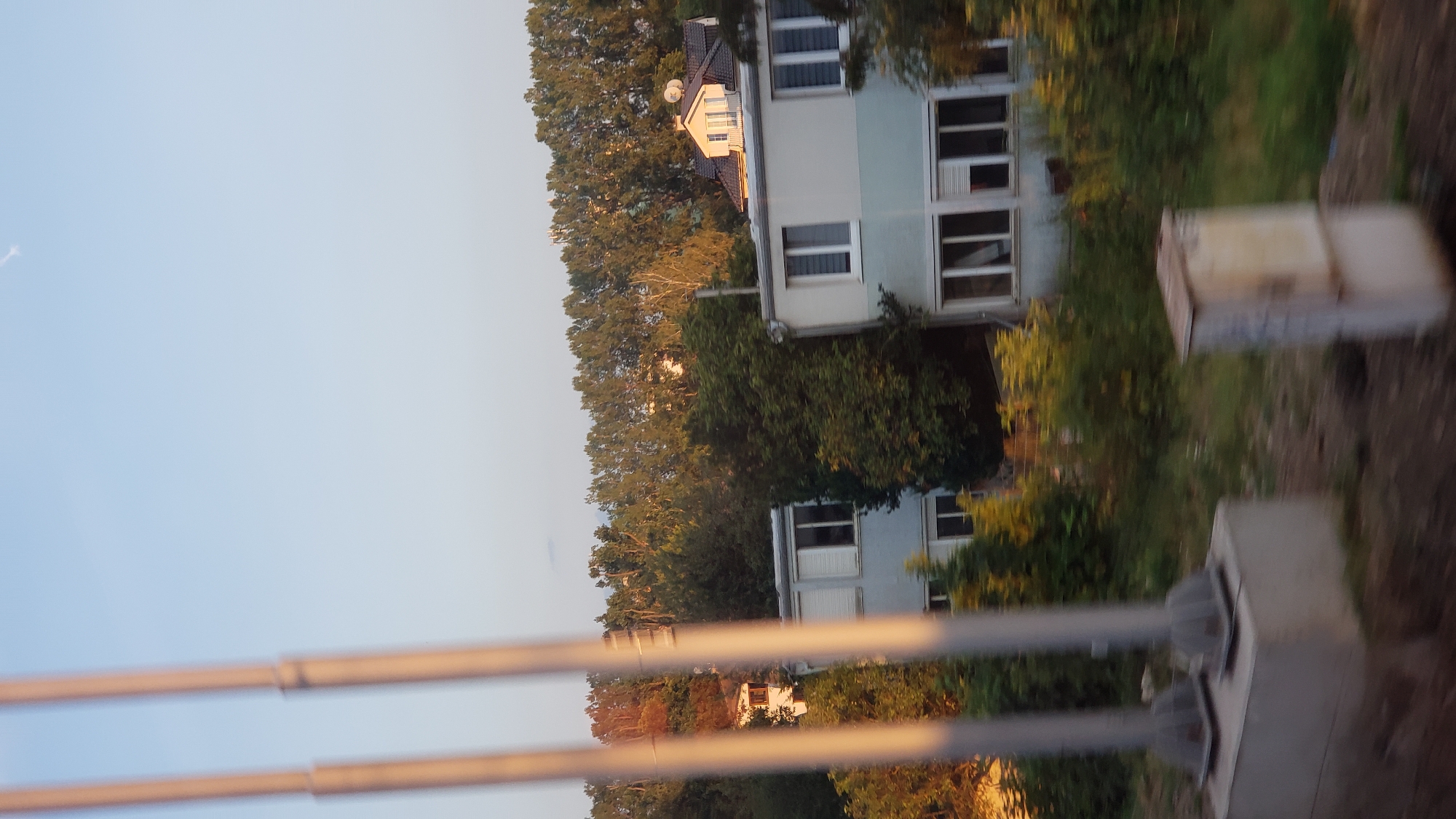 The image, taken from a moving vehicle, shows a view of a residential area with trees and buildings. In the foreground, two tall, grey poles with conical bases are visible on a concrete platform, suggesting they might be part of a lighting or utility system. To the right of these poles, a small, rectangular, light-colored utility box with visible graffiti is present.
Behind the poles, two modern, multi-story buildings are partially obscured by greenery. The building on the left has white-framed windows and what appear to be rolled-down blinds. The building on the right is a lighter blue and grey structure with multiple windows, some of which also have blinds. Further in the background, a house with a darker roof and dormer windows is nestled among mature trees. A dense line of trees forms the backdrop, with the sky above being a clear, light blue with a faint contrail visible.
The overall scene suggests an outdoor environment, possibly a suburban or semi-rural setting within the city of Poprad, Slovakia, given the prompt's context. The lighting indicates it is likely late afternoon or early evening, with the sun casting a warm glow on the trees. There are no people or animals visible in the frame.