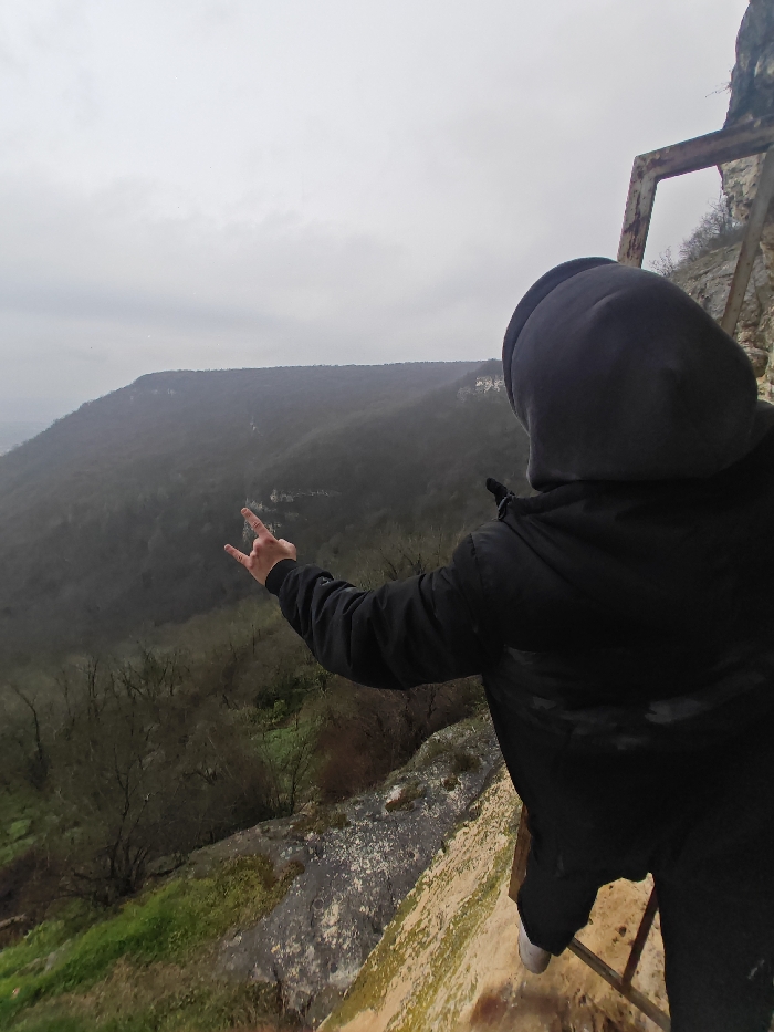 An individual, viewed from the back, stands on a narrow, moss-covered rock ledge overlooking a densely forested valley under an overcast sky. The location is the Shumen Plateau, near Shumen, Bulgaria. The subject is wearing a dark hooded jacket with the hood up, dark pants, and light-colored footwear. Their left arm is extended forward and slightly to the left, with the hand forming the "rock on" or "devil horns" gesture. A rusty, square metal frame structure is partially visible to the immediate right of the individual, attached to the vertical rock face. The immediate foreground shows the texture of the rock ledge, while the midground descends into a steep slope covered with deciduous trees, appearing sparsely foliated. Distant forested hills are visible on the horizon beneath a uniform gray sky.