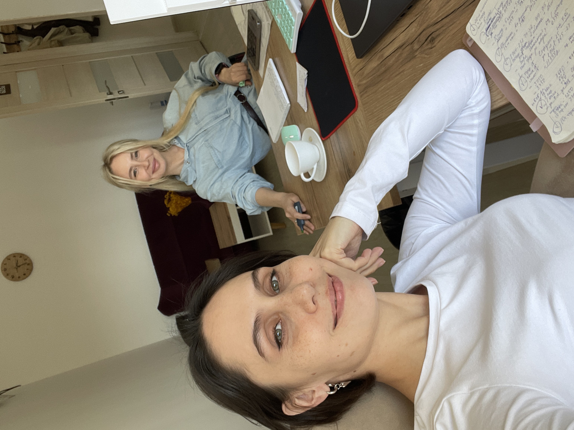 Two women are at a wooden desk. One woman with dark hair is taking a selfie, wearing a white shirt. The other woman with blonde hair is wearing a denim shirt. There is a white cup on a saucer, an electronic tablet, a keyboard, and a notepad. A clock on the wall can be seen in the background, along with a maroon couch. The OCR mentions figures related to finances, which are written on the notepad. No location context is given.