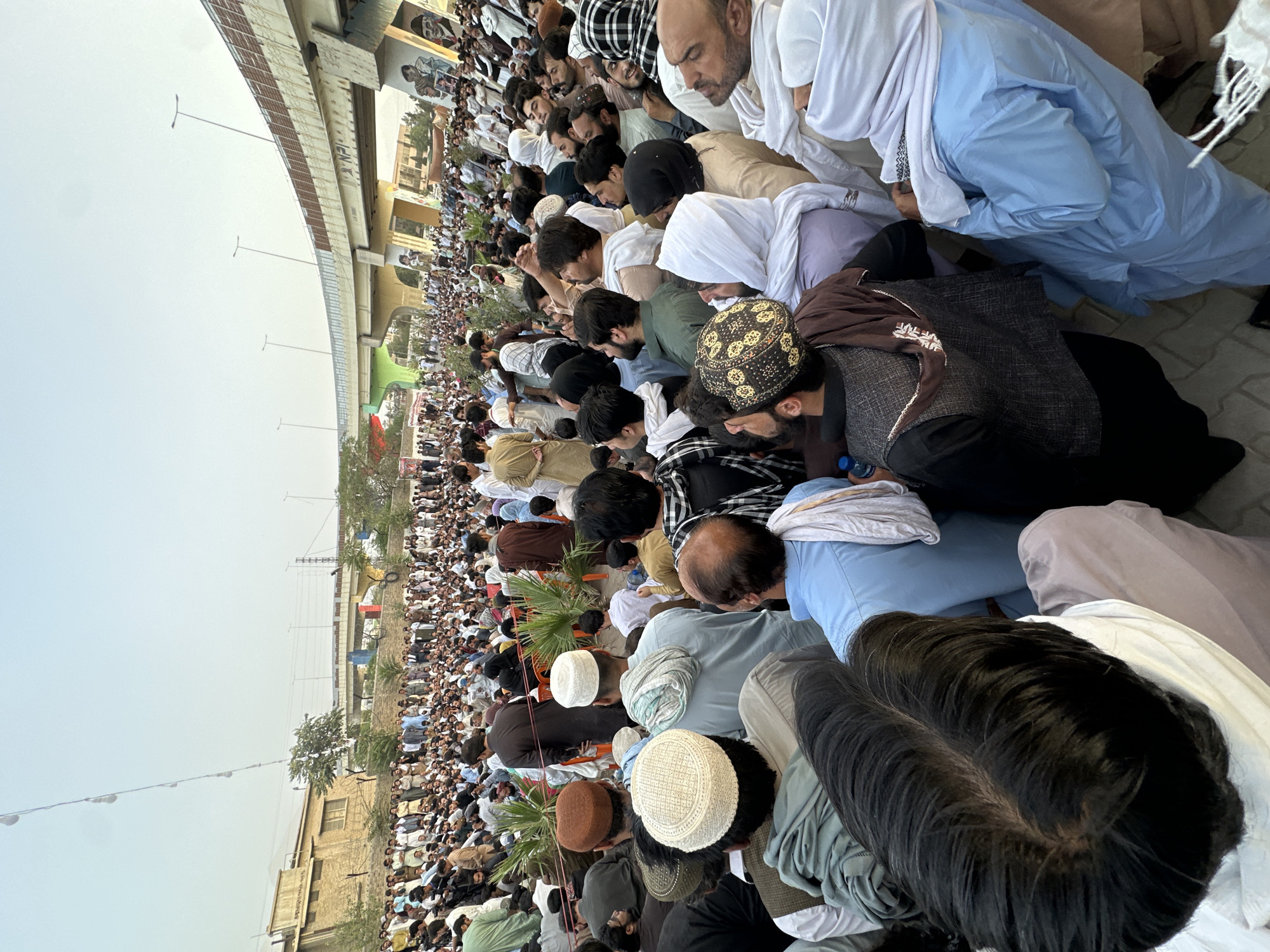 A wide shot captures a large gathering of people in Quetta City Tehsil, Pakistan, under a cloudy sky. The crowd, densely packed, spans across a paved area that appears to be an outdoor public space. A prominent overpass, painted with colorful murals featuring portraits, curves above the assembly. The people, predominantly men, are dressed in traditional Pakistani attire, including shalwar kameez and various types of caps and turbans. Many are looking in the same direction, suggesting they are attending an event or listening to a speaker. In the background, beyond the overpass, there are hints of buildings and trees. The overall scene conveys a sense of community and collective participation in an outdoor event.
