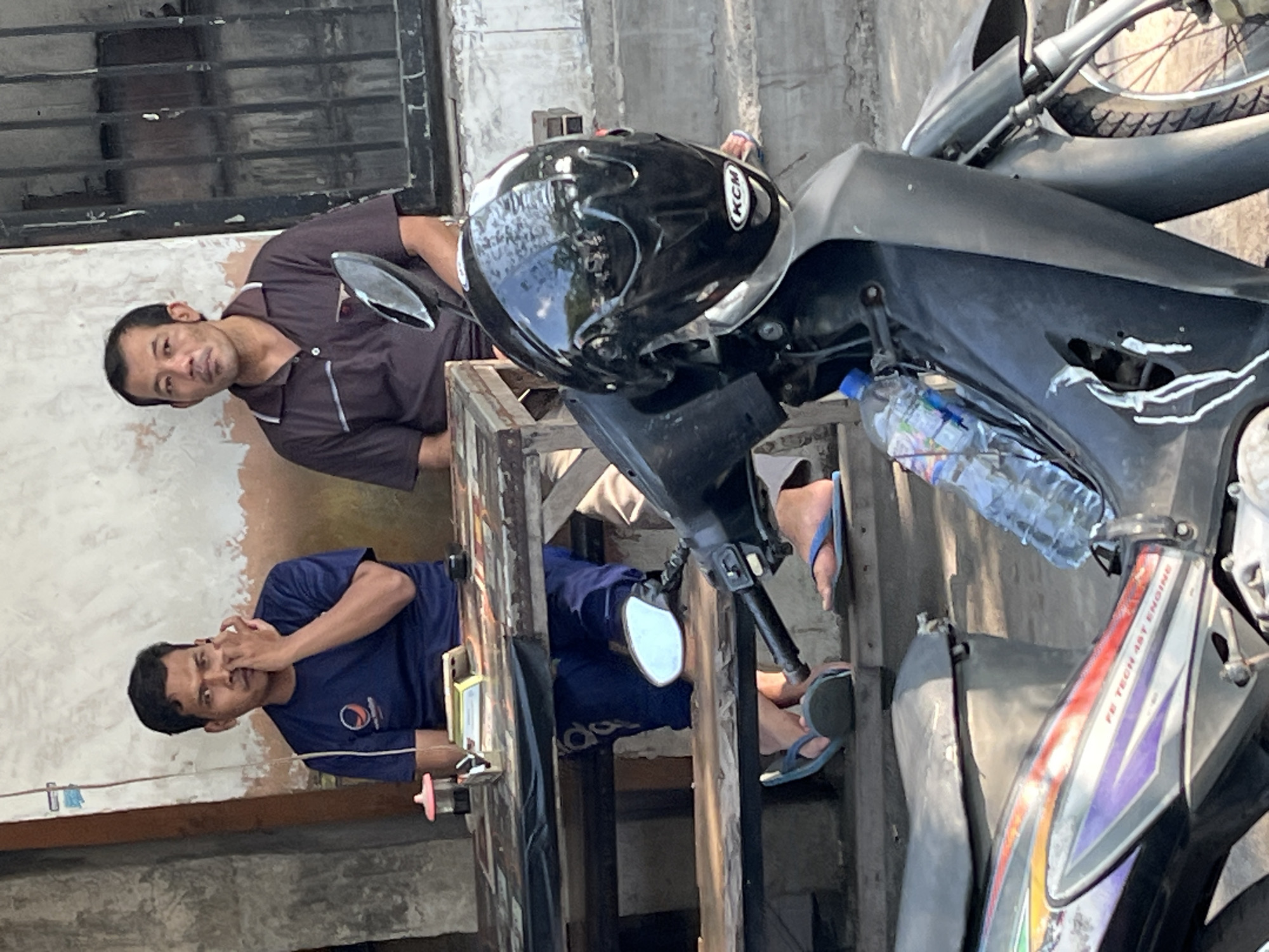 In Kludan, Indonesia, two men are seated at a wooden table outdoors, with a motorcycle in the foreground. The man on the left, wearing a navy blue t-shirt with a logo, is touching his nose with his right hand. The man on the right, in a brown polo shirt, looks towards the viewer. A black motorcycle helmet rests on the motorcycle's front. The environment appears to be a street or an open-air seating area, with a weathered wall and barred windows visible in the background.