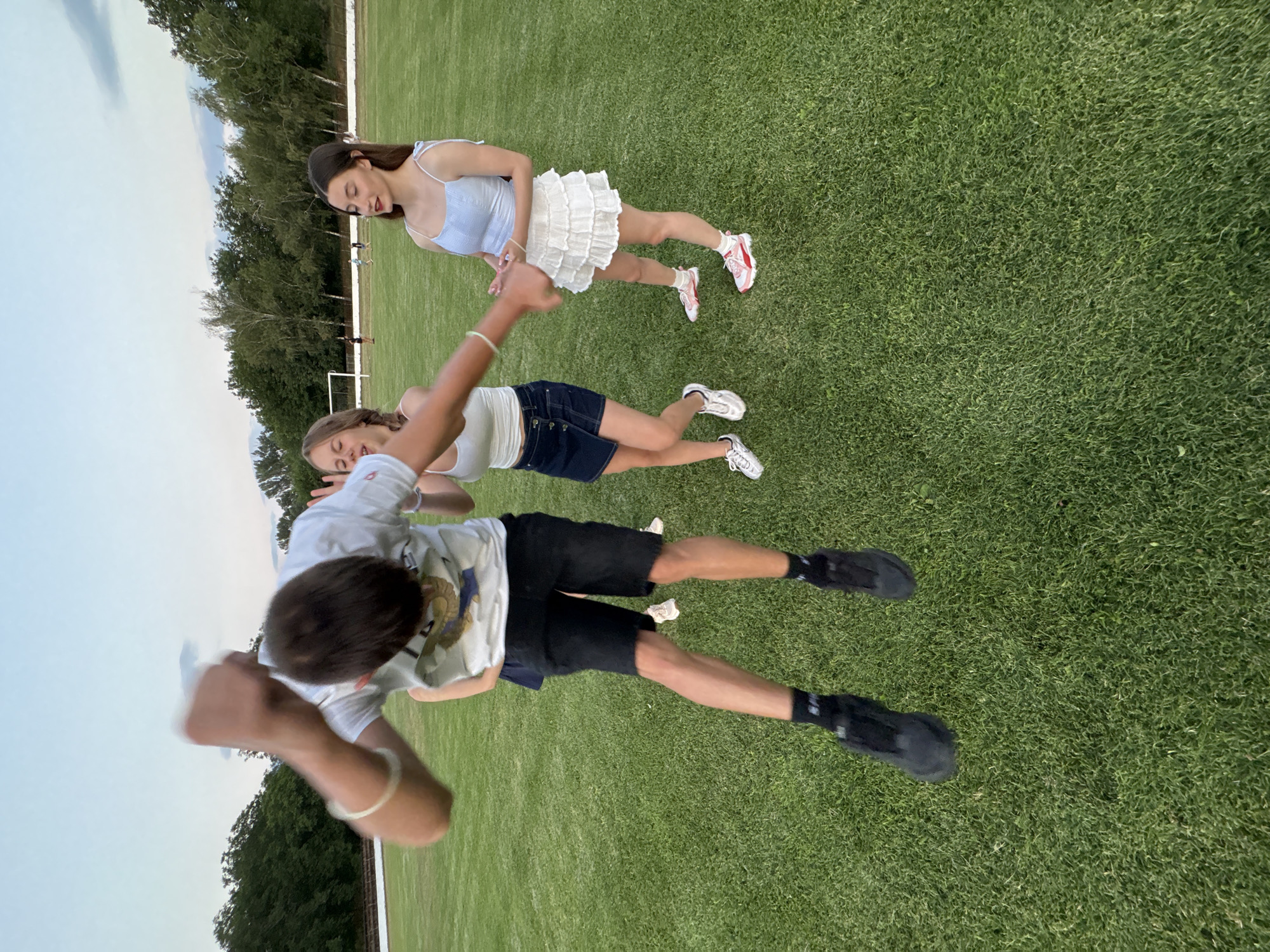 The image shows a group of four young people on a grassy field, likely a sports field, in Boboshevo, Bulgaria. In the foreground, a young male is captured mid-air, jumping with his arms raised and fists clenched. He is wearing a white t-shirt with a graphic on the front, black shorts, black socks, and black shoes. To his right, a young female is looking away from the camera with her eyes closed and mouth open, appearing to be reacting to the jumper. She is wearing a white tank top and a dark blue denim skirt. To the right of her, another young female stands facing the camera, wearing a light blue crop top and a white ruffled skirt, with pink and white sneakers. In the background, a soccer goal is visible, and a few other people are scattered further away on the field. The sky is overcast with a gradient from light blue to darker gray, suggesting it might be late afternoon or early evening. The environment is an outdoor grassy area with trees lining the perimeter.