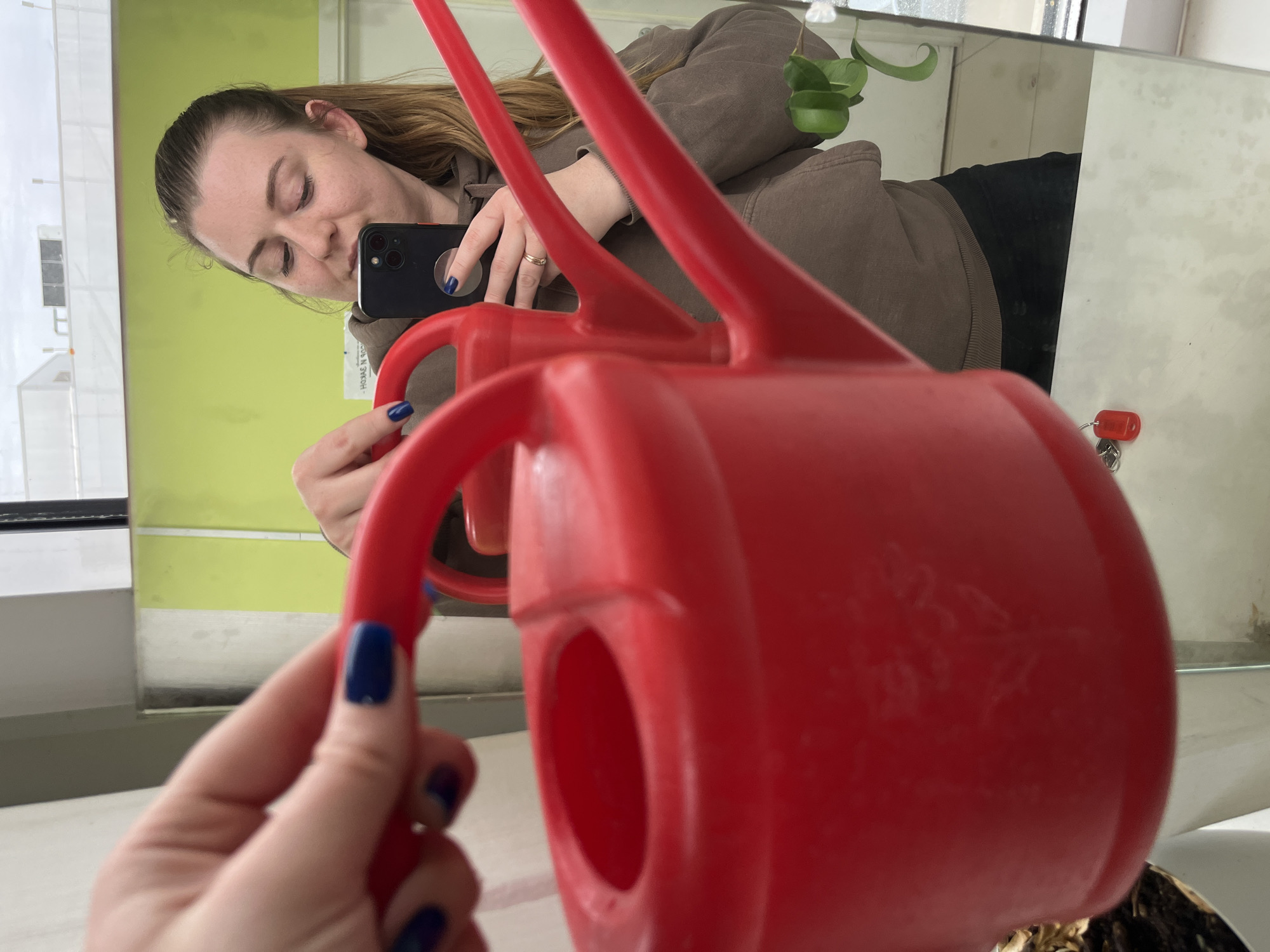 A woman is taking a selfie in a mirror while holding a red watering can. The woman is wearing a brown sweatshirt and has dark hair pulled back. She is holding a black smartphone. The mirror reflects the woman and the surrounding environment. A green plant is visible in the upper part of the mirror. The reflection shows the woman is in a room with a light green wall. A white counter is below the mirror. The lower part of the watering can sits near a plant with dark dry leaves. There is no indication in the picture this image was taken in Klin, Russia.