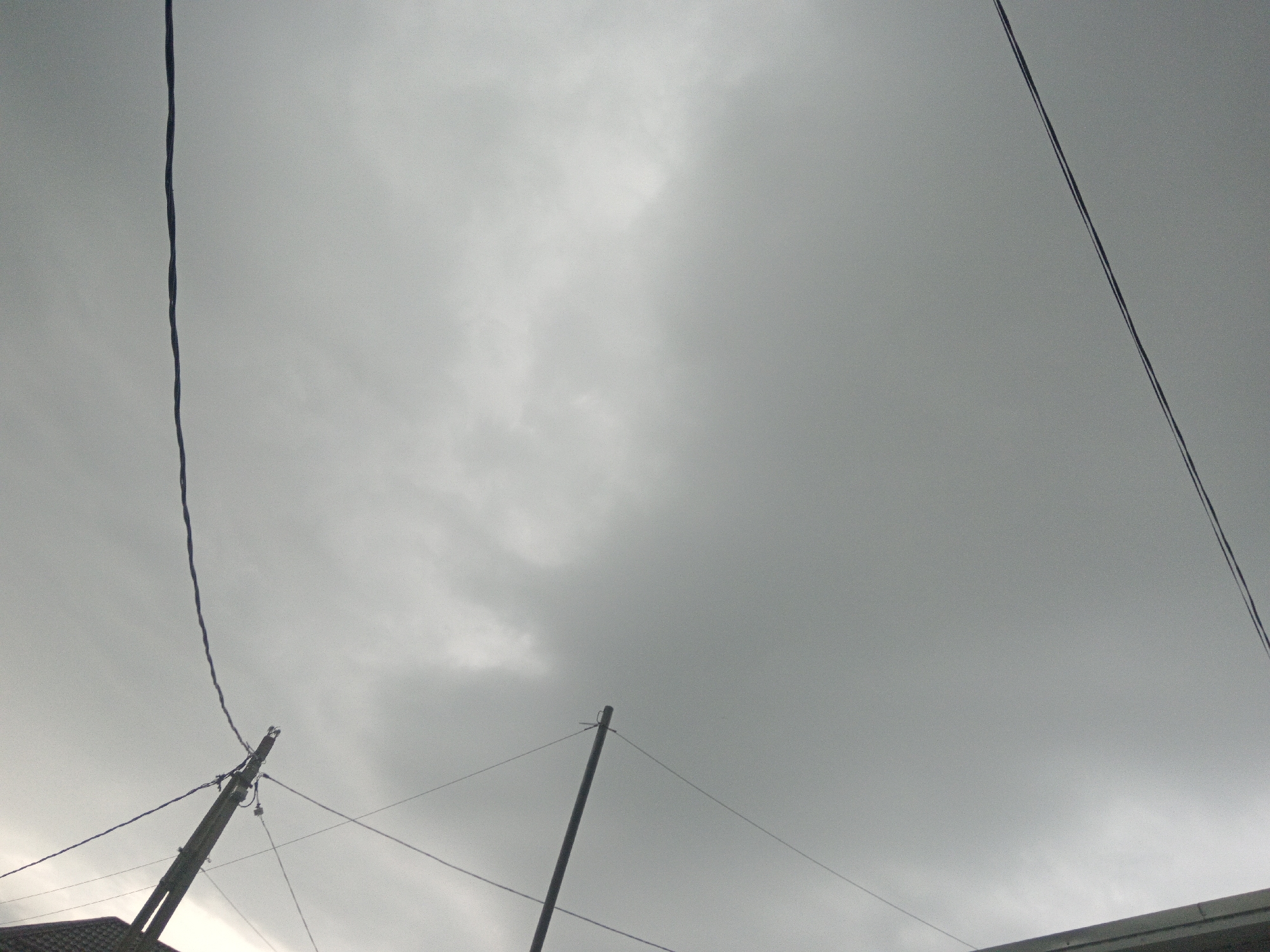 The image depicts an overcast sky above a portion of a building's roof in Uchkeken, Russia. Two utility poles are visible, one on the left and one in the lower center, with wires extending between them and across the frame. A dark, thin wire hangs vertically on the left side of the image, and another thicker, dark wire runs diagonally from the upper right towards the center. The sky is predominantly gray with some lighter, cloud-like formations. The building roof on the bottom edge appears dark and textured. There are no people or animals visible in the image. The scene conveys a utilitarian, outdoor environment.