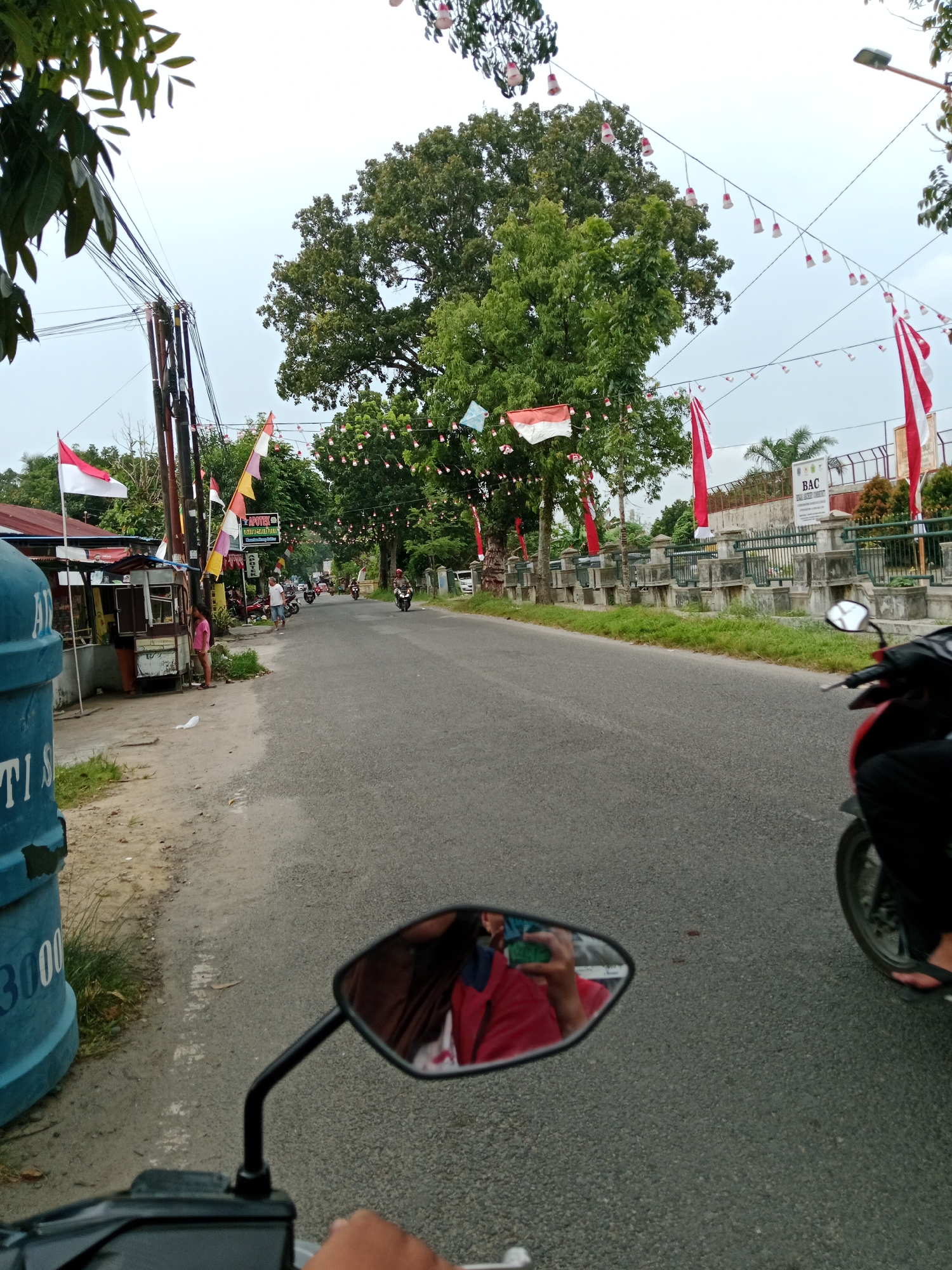 The image depicts a street scene in the City of Binjai, Indonesia, likely during a festive occasion, given the abundance of Indonesian flags (red and white) and decorative lights strung across the road. In the foreground, a motorcycle handlebar and mirror are visible, indicating the image was taken from the perspective of a rider. The mirror reflects the rider, who is wearing a red shirt, and appears to be taking a photograph.
To the left of the road, a blue cylindrical object with white lettering, possibly a water tank or large container, is present. Adjacent to it are small roadside stalls or shops, one with a red roof. A young girl in a pink shirt stands near these stalls.
The road itself is paved and appears to be a two-lane street with moderate traffic, including several motorcycles. People are seen walking and standing along the roadside. In the background, there is a row of trees lining the street, and beyond that, a building with a fence and a sign that reads "BAC." The sky is overcast. The presence of Indonesian flags suggests the photo may have been taken around Independence Day (August 17th) or another national celebration.