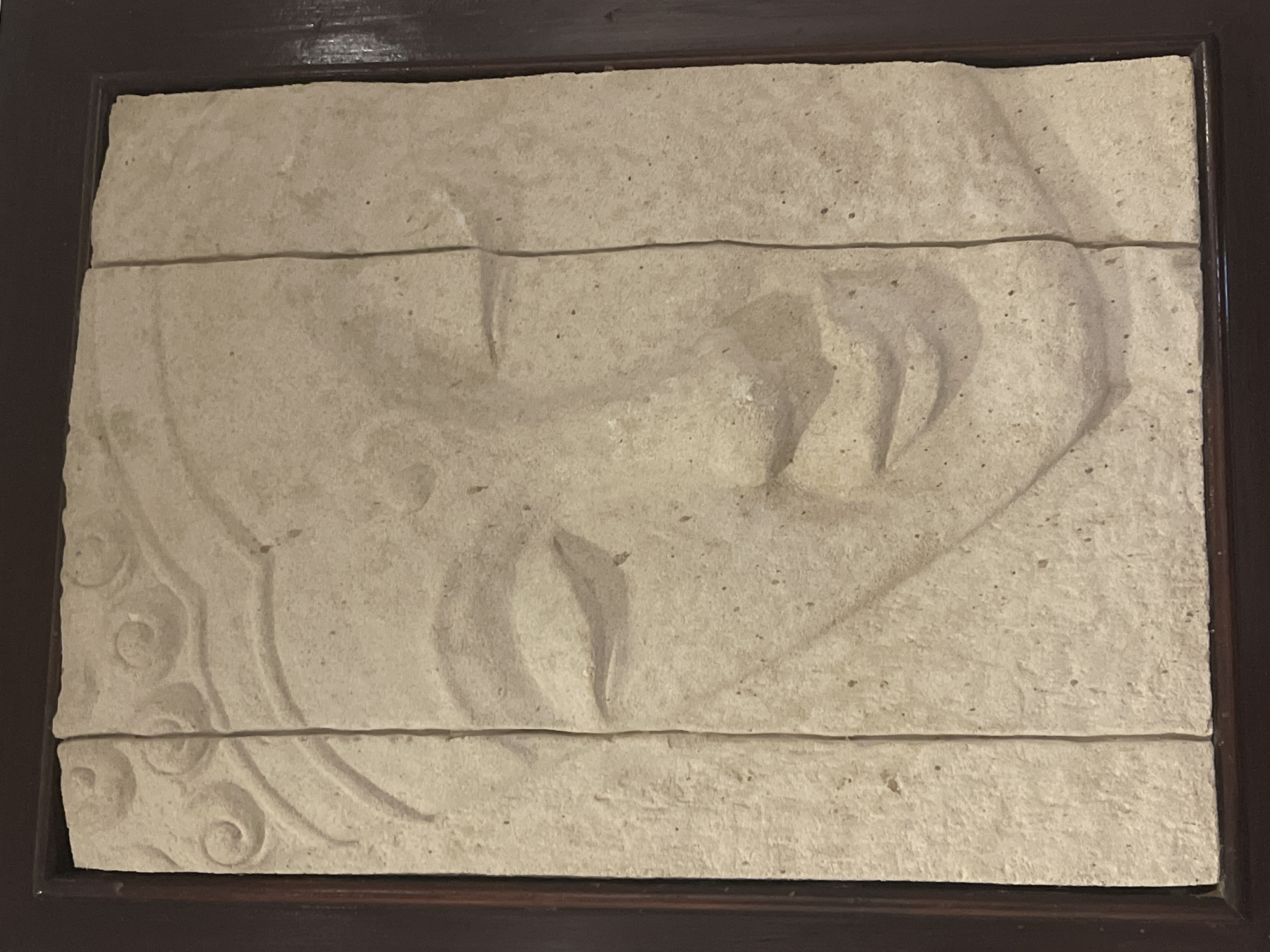 The image displays a light-colored stone relief carving set within a dark brown, possibly wooden, frame. The carving predominantly features a profile view of a face, likely human, with a serene or sleeping expression, characterized by a prominent nose, lips, and what appears to be a closed eye or a defined eyebrow line. The facial features are subtly sculpted in low relief.
On the lower-left side of the carving, intricate swirling and wave-like ornamental patterns are visible, suggesting decorative elements such as hair, a stylized headdress, or surrounding iconography. The material of the carving is porous and textured, resembling sandstone or a similar type of aged, light-colored stone, with small dark specks indicative of its natural composition or wear.
The entire relief is segmented horizontally into at least three distinct sections by two noticeable straight lines running across its width, indicating that the carving is composed of multiple stone slabs fitted together. The dark brown frame provides a contrasting border to the light stone artwork. The lighting highlights the three-dimensional contours of the relief, creating shadows that define the depth and details of the carving. There is no activity taking place, as it is a static art piece, and no visible text is present.