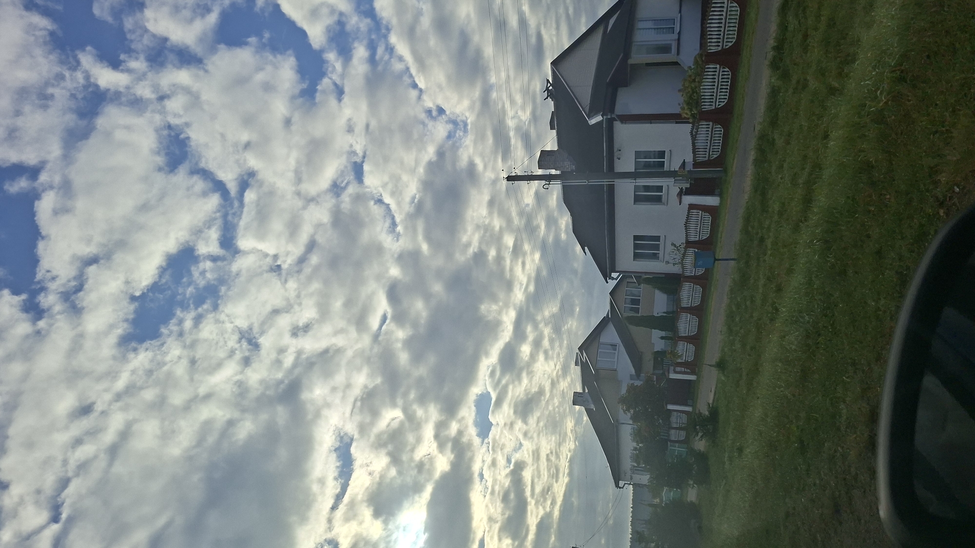 The image depicts a street scene in Парафʼянаўскі сельскі Савет, Belarus, under a cloudy sky. Two houses are visible, with the one on the right being a white, two-story structure with a dark roof and several windows. The house on the left is partially obscured but appears to be a similar style. Both houses are bordered by a decorative fence with a dark base and white arched elements. A tall utility pole with wires stands near the house on the right. The foreground features a grassy area with some trees, and a portion of a dark-colored vehicle is visible at the bottom. The sky is filled with a mixture of white and gray clouds, with patches of blue sky showing through.