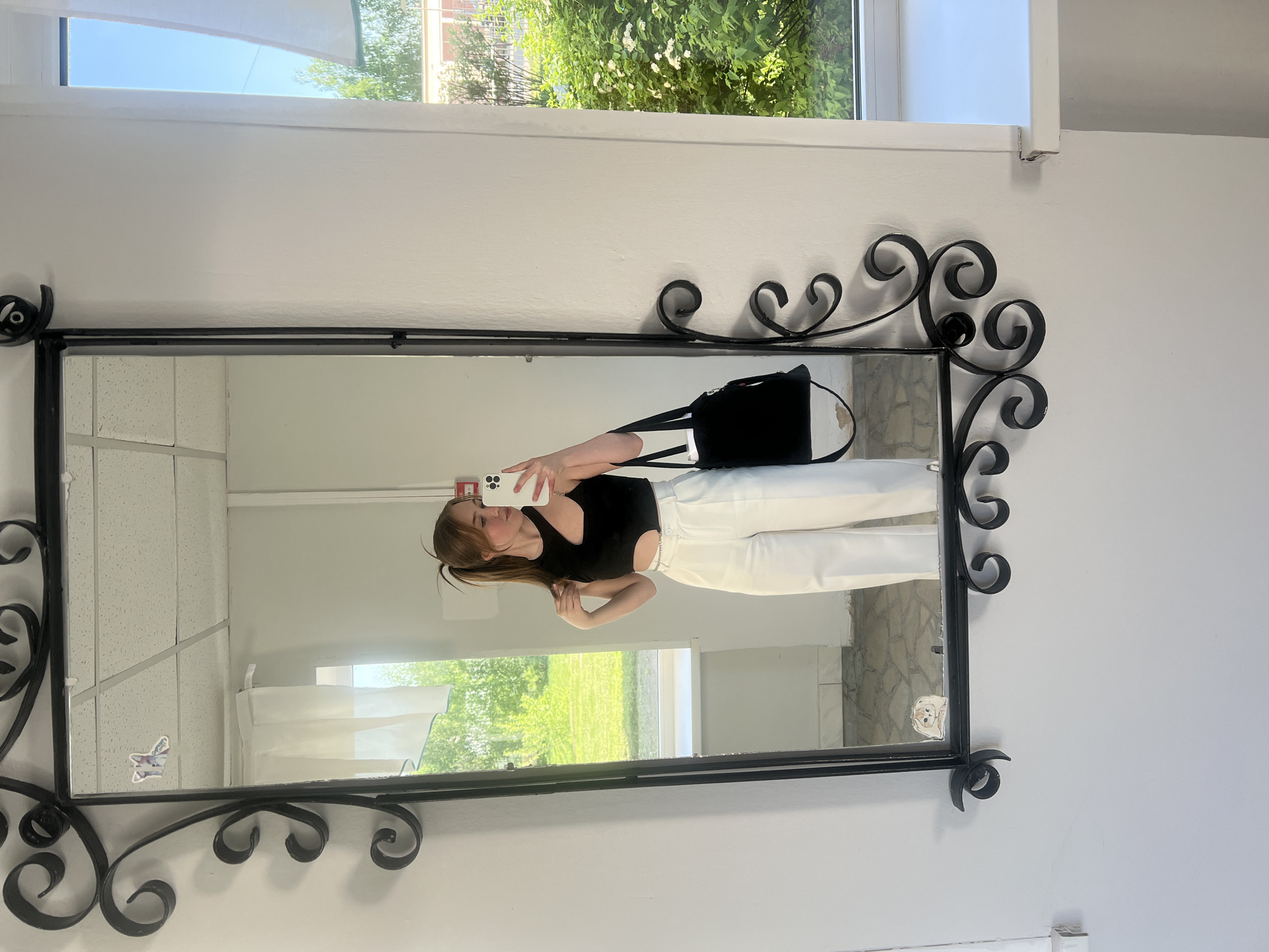 The image shows a young woman taking a selfie in a mirror. The mirror is ornate with a dark, curly metal frame. The woman is wearing a black top and white pants. She is holding a smartphone in her right hand, and it is pointed towards the mirror. Her left arm is bent at the elbow, and her hand is near her shoulder. A black bag with white straps hangs over her shoulder. The background reflected in the mirror shows a room with white walls, windows, and a view of greenery outside. There is also a sticker on the mirror. The location is inferred to be Chaikovsky, Russia, but without further context, this is not verifiable from the image alone.