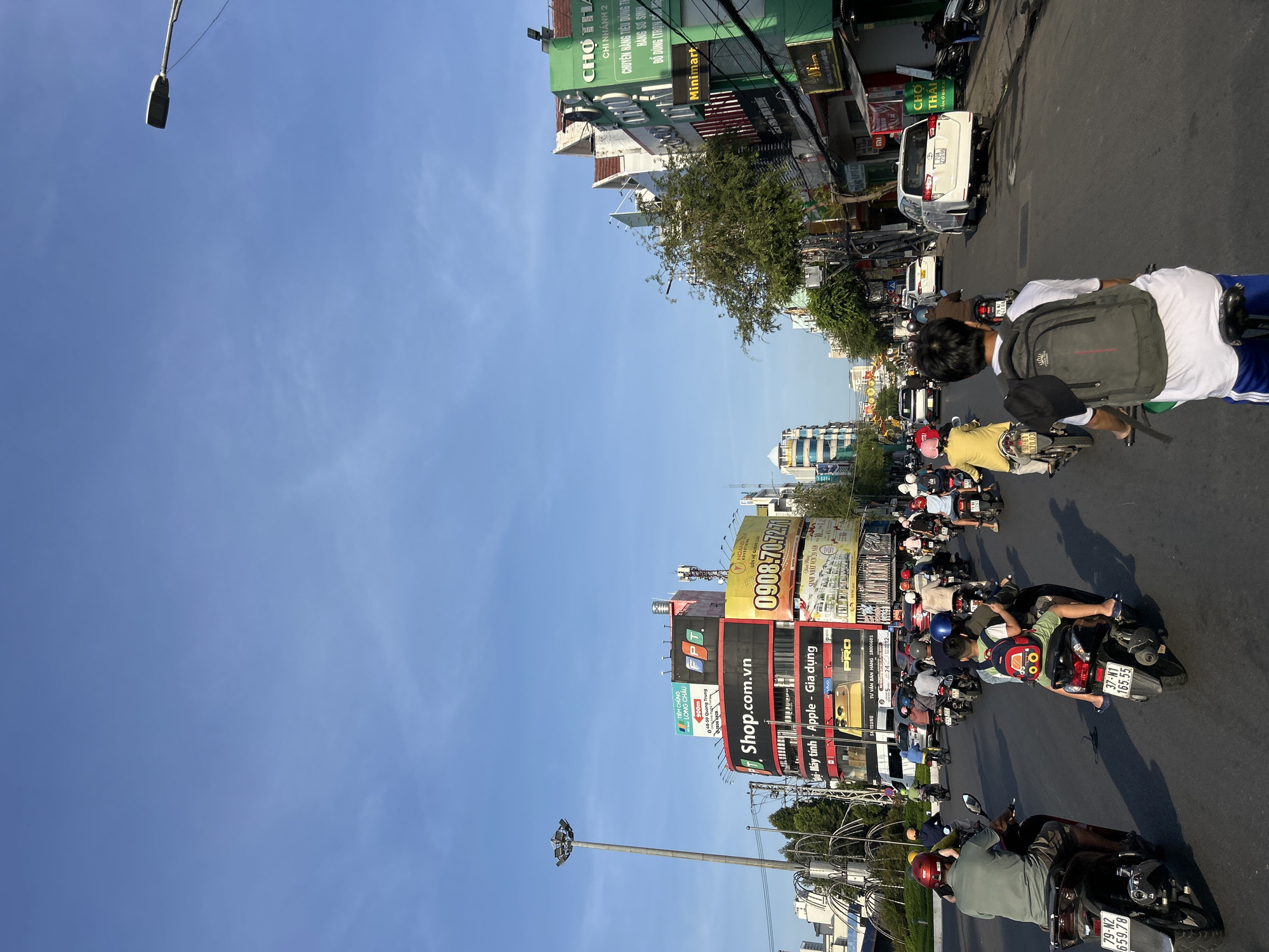 This is a vertical, eye-level shot of a busy street scene in Nha Trang Ward, Vietnam. The sky is a clear, bright blue with minimal clouds. A tall street lamp stands on the left side of the frame. The road is filled with a multitude of motorcycles and scooters carrying passengers, indicating a common mode of transportation. The buildings lining the street are colorful and feature numerous advertisements and signs, typical of commercial areas. Some buildings have multiple stories, and some appear to be shops or businesses. The overall atmosphere is one of active urban life.
On the street, several people are riding motorcycles. In the foreground, a person wearing a green shirt and a red helmet is riding a dark-colored motorcycle. To their right, another person wearing a yellow jacket and a white shirt is riding a motorcycle, with a child in a yellow shirt and a red backpack on the back. Further ahead, more motorcycles and people are visible, some with helmets and some without. Vehicles are moving in the same direction.
The buildings display a variety of commercial signage. One prominent sign reads "Shop.com.vn" with "Apple - Gia dụng" and "vivo" below it. Another large yellow sign features the phone number "0908.70.72.71". A green building on the right has signs that include "CHỢ THÁI" and "Minimart". License plates are visible on some of the motorcycles, such as "79-N2 659.78" and "37-N1 165.55". A white car, possibly a minivan, is parked on the right side of the road. Electrical wires are strung across the street, and trees are present along the sidewalk. The sunlight casts shadows on the road, suggesting it is daytime, likely morning or afternoon.