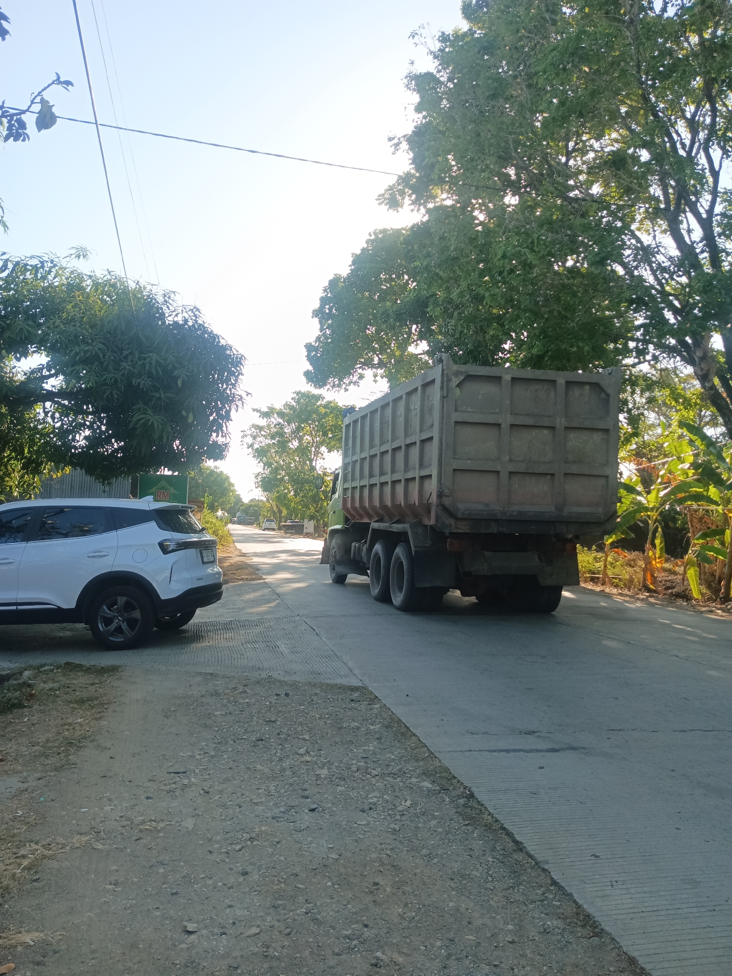The image shows a road in the city of Bowong Cindea, Indonesia, with a dump truck driving away from the camera on the right side of the road. A white SUV is parked on the left side of the road, facing away from the camera. The road is paved with concrete and has a gravel shoulder. There are trees and vegetation on both sides of the road, and the sky is clear and blue. The sun is shining brightly, casting shadows on the road. The dump truck appears to be empty.