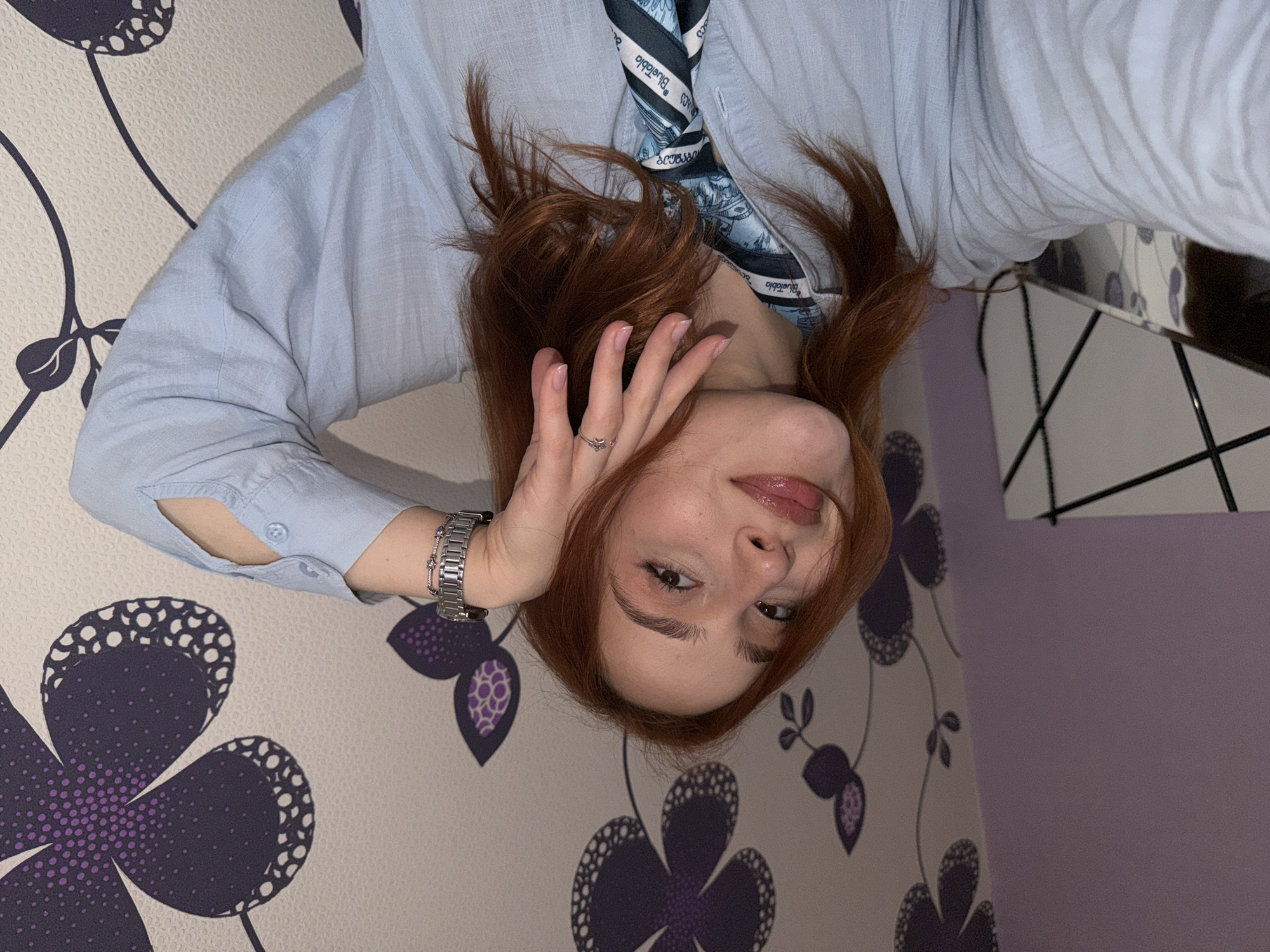A young woman with reddish-brown hair is positioned upside down in the frame. She is wearing a light blue button-down shirt with a blue patterned scarf. A silver wristwatch is visible on her left wrist, and a ring is on her finger. Her hand is near her face, and she is partially smiling. The background consists of a patterned wallpaper with large purple and white floral designs on one side and a plain purple wall with a partially visible modern lamp with black lines on the other. The location information is unavailable.