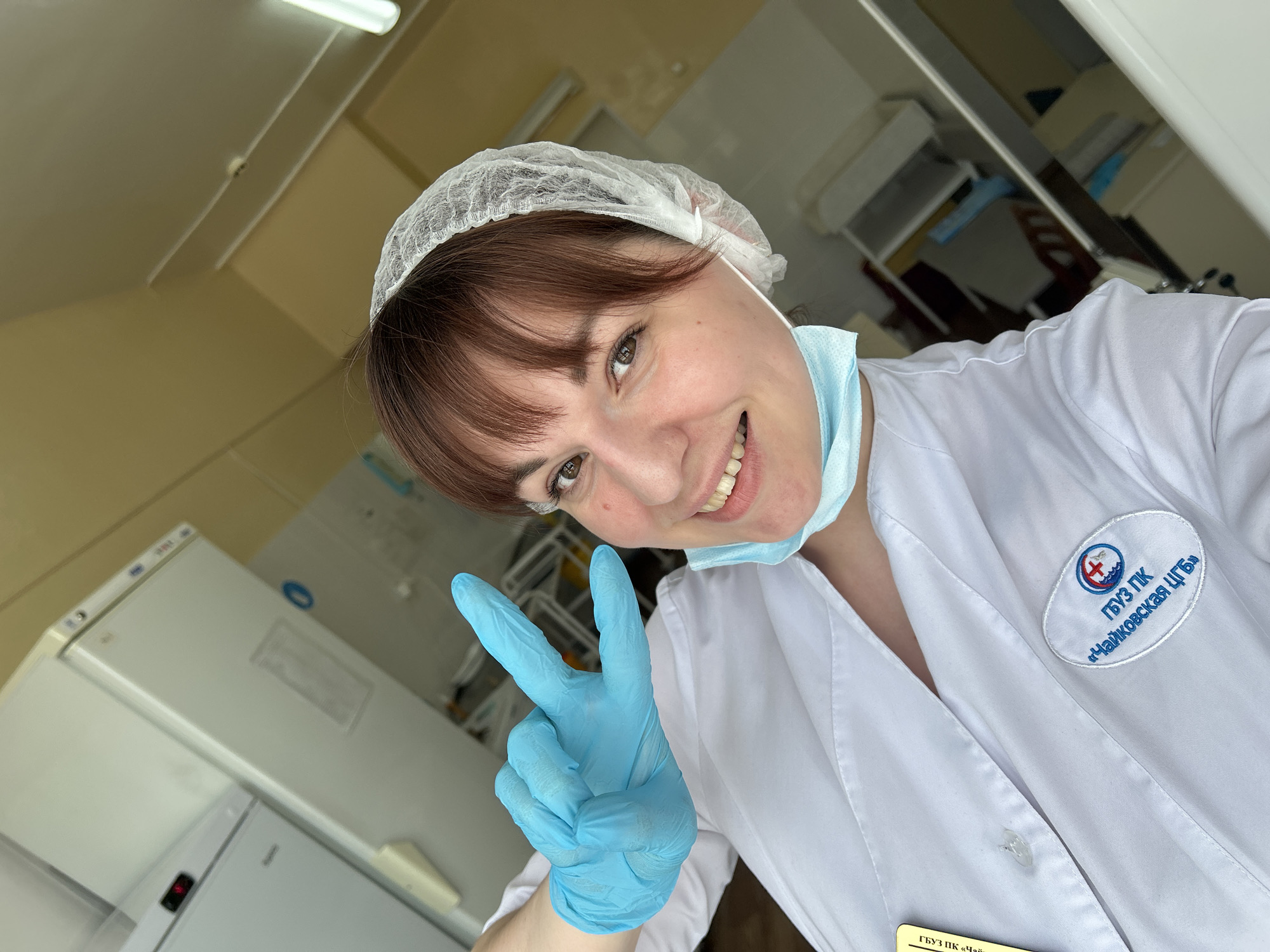 A female individual, appearing to be in a clinical or laboratory setting, is depicted in a selfie. She has brown hair with bangs, brown eyes, and is smiling directly at the camera. She wears a white lab coat, a disposable white hair net, and a light blue disposable surgical mask pulled down around her chin. Her left hand, covered by a light blue disposable glove, is raised, forming a "peace" or "victory" sign with the index and middle fingers extended. On the left chest area of her lab coat, an oval embroidered patch is visible, featuring a logo with waves, a medical cross, and a building, along with Cyrillic text reading "ГБУЗ ПК «Чайковская ЦГБ»," which translates to "State Budgetary Healthcare Institution of Perm Krai 'Chaykovskaya Central District Hospital'."
The background consists of light yellow walls. To the left and behind the individual, two white refrigerators or freezers are present; one has a red digital display. Fluorescent tube lighting is visible on the ceiling, illuminating the scene. Based on the uniform's insignia, the location is Chaykovskaya Central District Hospital, located in Chaykovsky, Perm Krai, Russia.