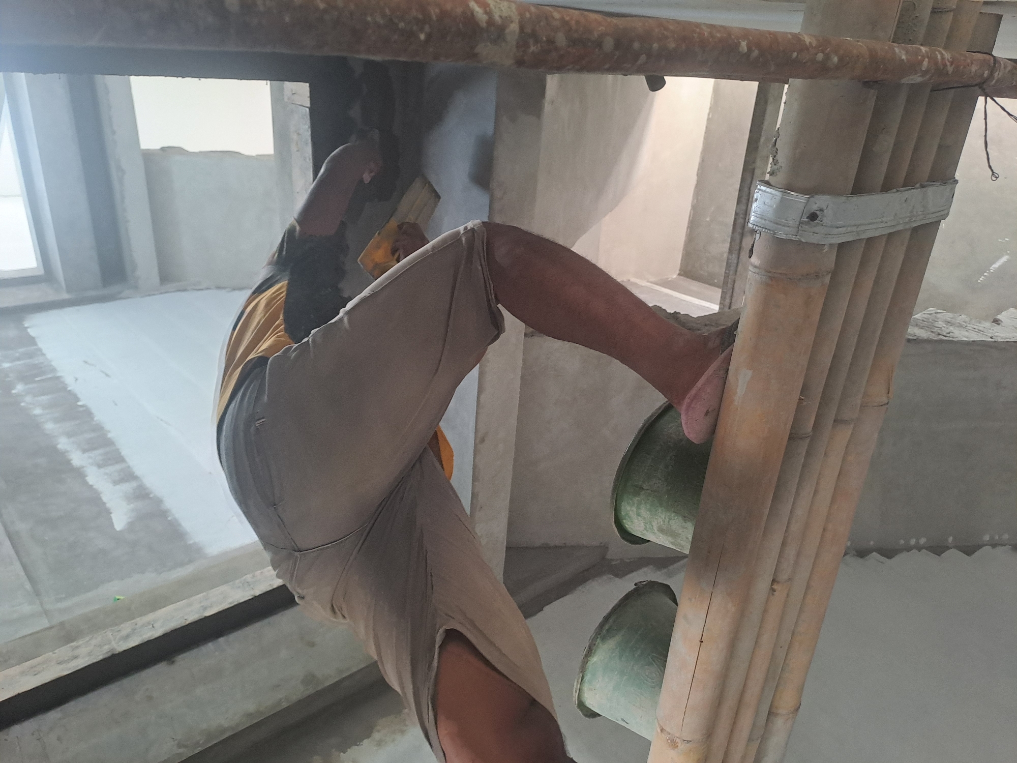 In Ngaglik, Indonesia, a construction worker is seen from a low angle, wearing tan shorts and a yellow and black t-shirt. They are standing on a bamboo scaffolding, applying plaster to a concrete wall with a trowel. Two green buckets are positioned behind them on the scaffolding. The environment appears to be an unfinished building with exposed concrete and scaffolding.