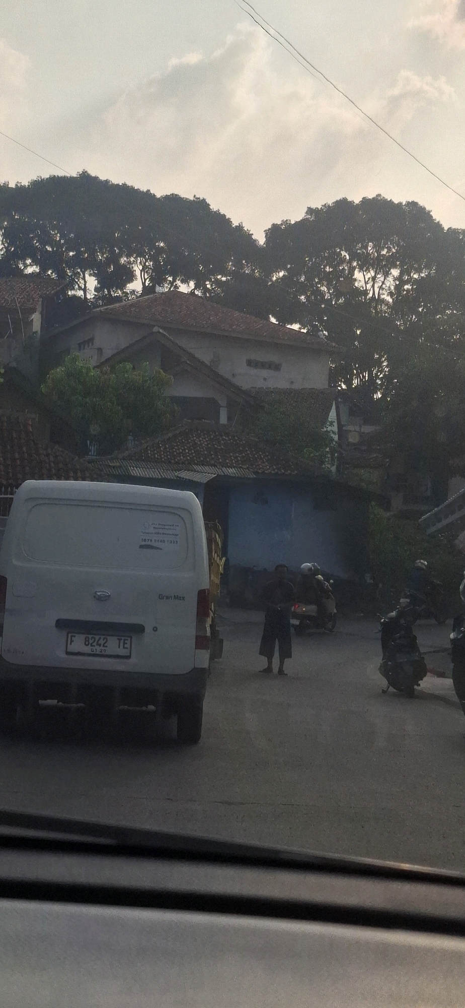 In the city of Cilaku, Indonesia, a street scene is captured under a hazy sky, likely during either dawn or dusk given the soft, diffused light. The foreground is dominated by the rear of a white van, identified by its license plate "F 8242 TE" and the "Gran Max" model designation. A portion of a yellow truck bed is visible attached to the van.
On the right side of the street, a man stands near a parked motorcycle. His attire consists of a dark top and light-colored, knee-length pants. Further down the road, a motorcycle with two occupants, one wearing a white helmet, is in motion. Another dark-colored motorcycle is parked on the right shoulder.
The background reveals a residential area characterized by houses with tiled roofs and lush trees that create a dense canopy. A power line is visible stretching across the upper part of the frame, indicating the presence of urban infrastructure. The overall impression is a quiet, possibly residential street in a tropical climate.