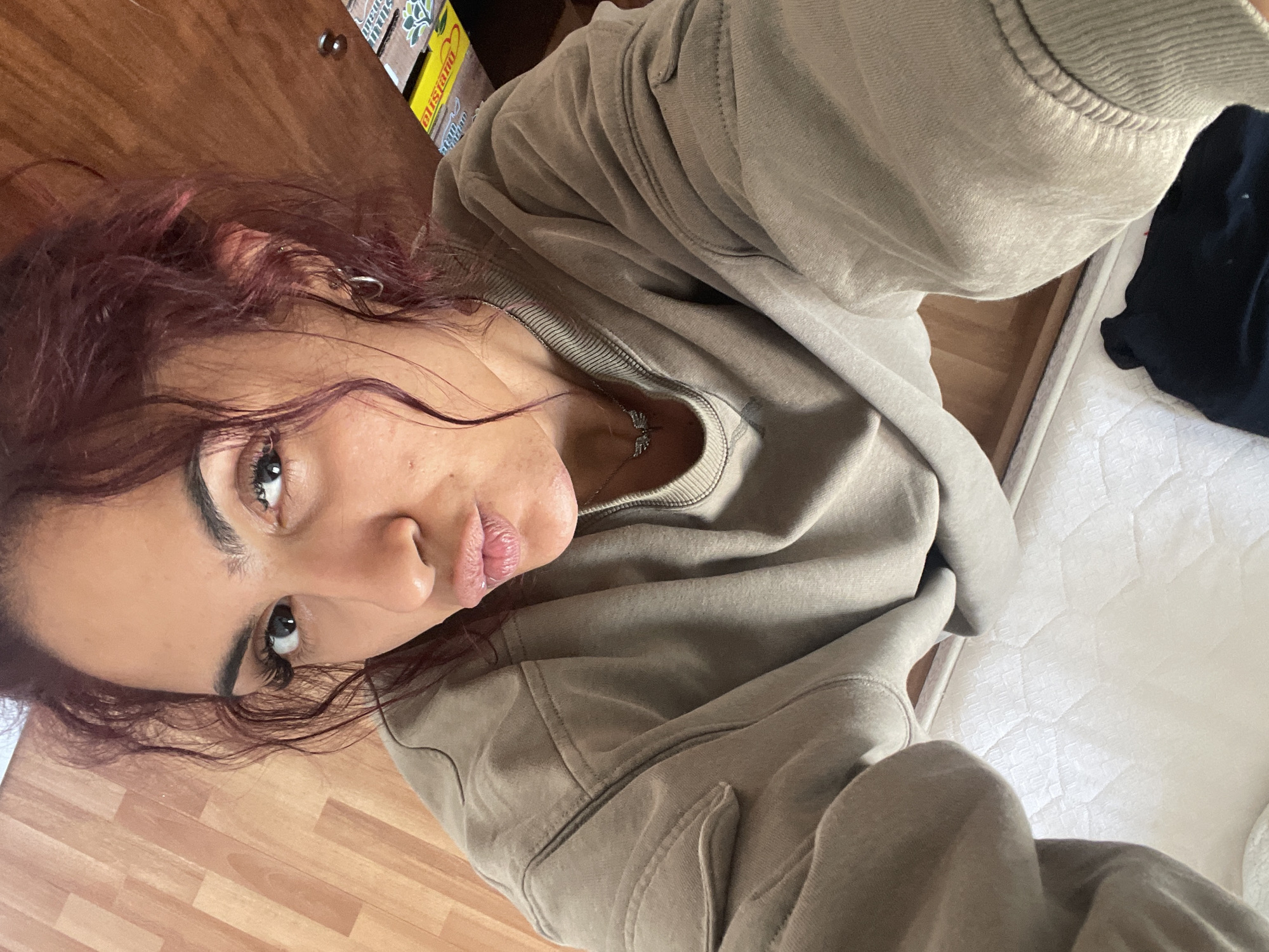 An image captured from a low-angle perspective displays a female individual, positioned centrally, with her head tilted slightly upwards. The subject has medium-dark skin tone, and her hair is curly, styled in a messy manner, exhibiting a reddish-brown coloration. She wears cosmetic eyelashes. Her lips are pursed in a "pout" expression, and her eyes gaze directly at the camera. A silver-toned hoop earring is visible in her right ear. Around her neck is a silver-toned necklace with a small winged pendant. She is dressed in a loose-fitting, long-sleeved, crew-neck sweatshirt of a light olive green or khaki hue, featuring stitched detailing on the shoulders and a decorative element on the upper left sleeve.
The background consists of two distinct sections. The upper portion, behind the subject's head, features a vertical wooden surface with light brown planking. In the upper right quadrant, an object or surface shows partial yellow and white text "DUBLIN" on a green and white background, accompanied by a leaf motif. The lower portion of the background, below the subject's shoulders, reveals a white quilted mattress or mattress cover with a diamond-patterned texture. A dark-colored fabric item is partially visible on the mattress in the lower right corner. The setting is an indoor environment in the city of Unknown, Unknown.