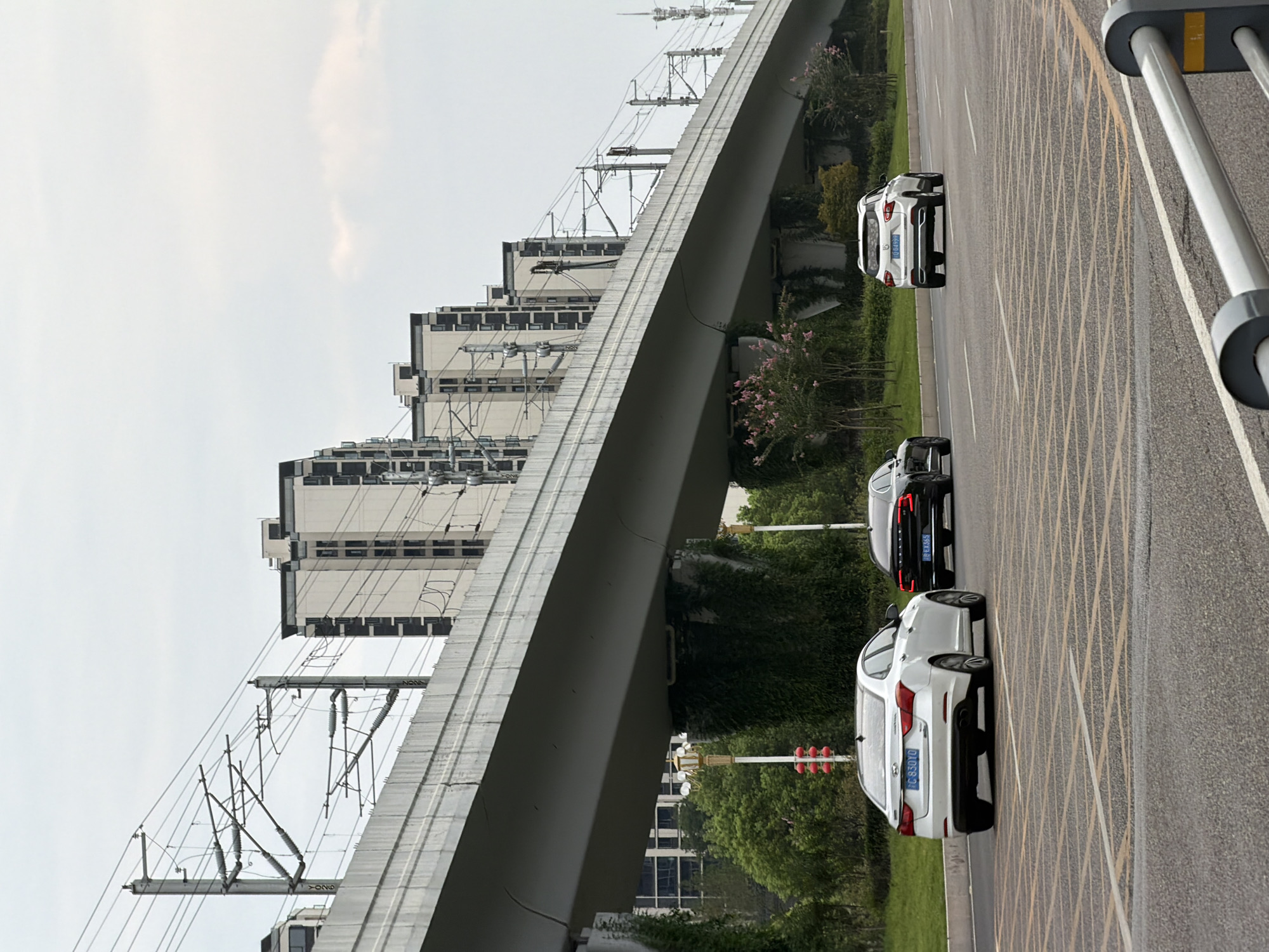 In Jiangjin, China, under an elevated overpass supporting an electrified railway line, a white sedan with license plate 'C. 830YO' travels on a multi-lane asphalt road. To its left, a black sedan is also on the road. Further ahead and to the right, a white SUV with license plate 'D.G4999' is also driving. Behind the overpass, a series of identical, multi-story apartment buildings with dark-framed windows are visible against a cloudy sky. The supports of the overpass are covered in green ivy, and decorative streetlights with red lanterns are present. The road has clear lane markings and crosswalks.