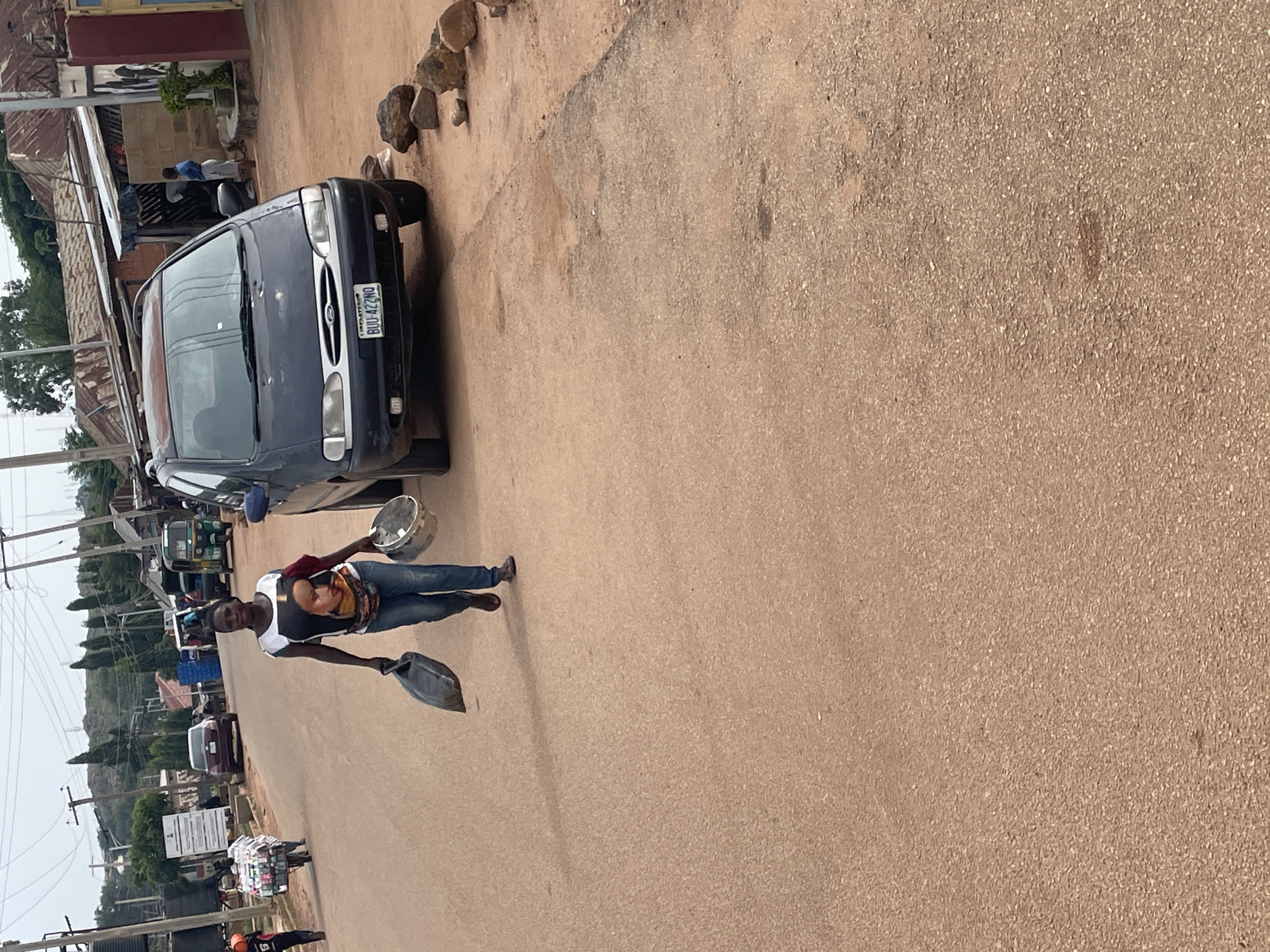 In the city of Jos, Nigeria, on a dirt road, a woman is walking, carrying a black bag in her left hand and a silver bucket in her right. She is wearing a white shirt with a picture of a woman's face on it, blue jeans, and red shoes. The woman is walking past a dark blue van with the license plate "BUU 422NO". In the background, there are several buildings with corrugated iron roofs, and a few trees. The sky is overcast.