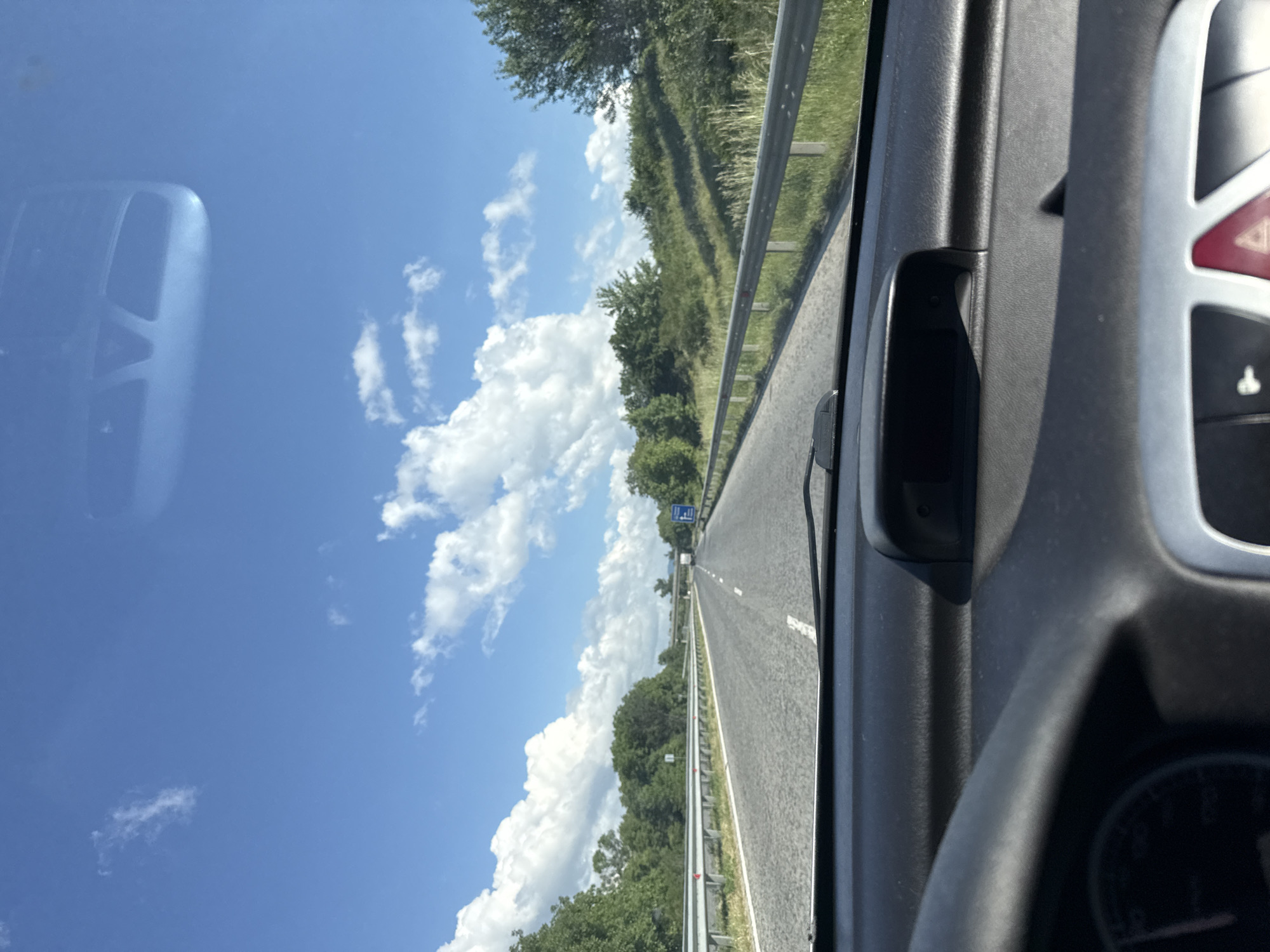 The image shows a view from inside a vehicle, likely a car, driving on a highway in the city of Zmeyovo, Bulgaria. The highway has two lanes and a metal guardrail. Lush green vegetation lines the side of the road. The sky is blue with fluffy white clouds. A sign is visible in the distance. The interior of the car shows part of the dashboard, a rearview mirror, and the steering wheel.