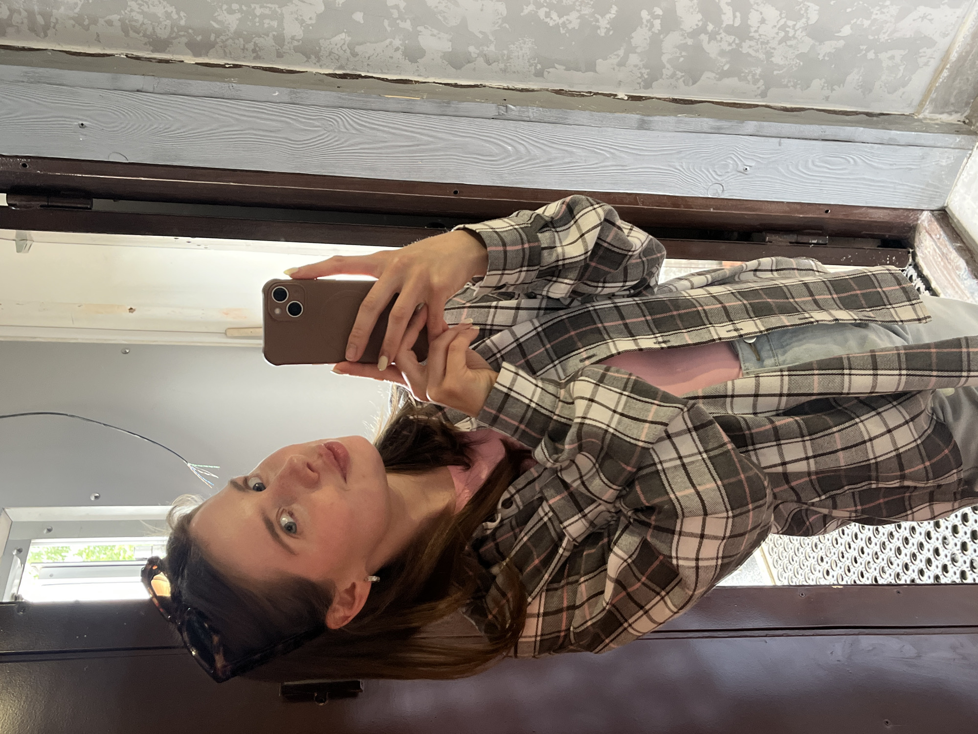 A young Caucasian woman with light brown hair, wearing sunglasses perched on her head, a pink t-shirt, and a grey, white, and pink plaid flannel shirt, stands in what appears to be an entryway. She is taking a selfie with her phone, which is held in her right hand. Her left hand is partially visible near her phone. Her expression is playful, with her lips pursed. The background suggests an interior space with a dark brown door on the left, a partially visible metal grate floor, and a wall with a light-colored wood-paneled section and a textured grey section on the right. The lighting is bright, suggesting daylight. The scene does not provide specific landmarks to confirm a location within the city of Chaikovsky, Russia.