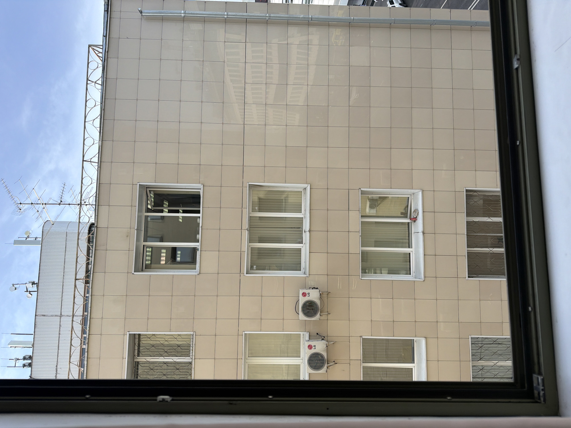 View from inside a building looking out at an adjacent building with a beige tiled facade. The building has windows arranged in a grid pattern, some with blinds. Two air conditioning units are mounted on the facade between two of the windows. To the left, part of another structure with an antenna and decorative railing is visible. The sky is partially visible in the upper left. The image is framed by a dark-colored window frame. The location is Moscow, Russia.