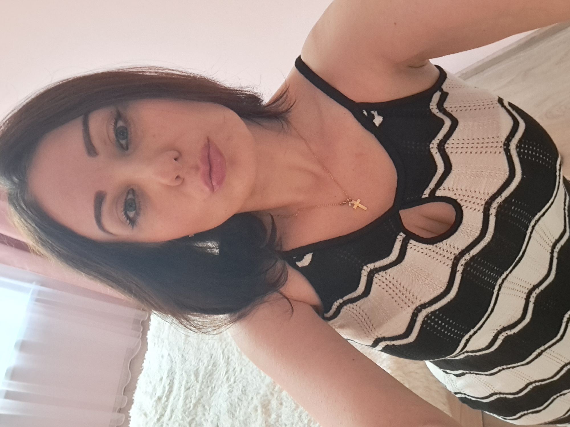 A woman is captured in a close-up, selfie-style photograph. She has medium-length dark brown hair, styled loosely around her face. Her eyes are wide open, and she is puckering her lips. She is wearing a black and white striped tank top with a cut-out detail at the neckline, and a gold cross necklace. The background suggests an indoor setting, with a soft, textured rug visible on the left and a plain pink wall on the right. A window with sheer curtains is also partially visible in the upper left corner. The lighting is soft and diffused. There is no indication of the location being Irkutsk, Russia.