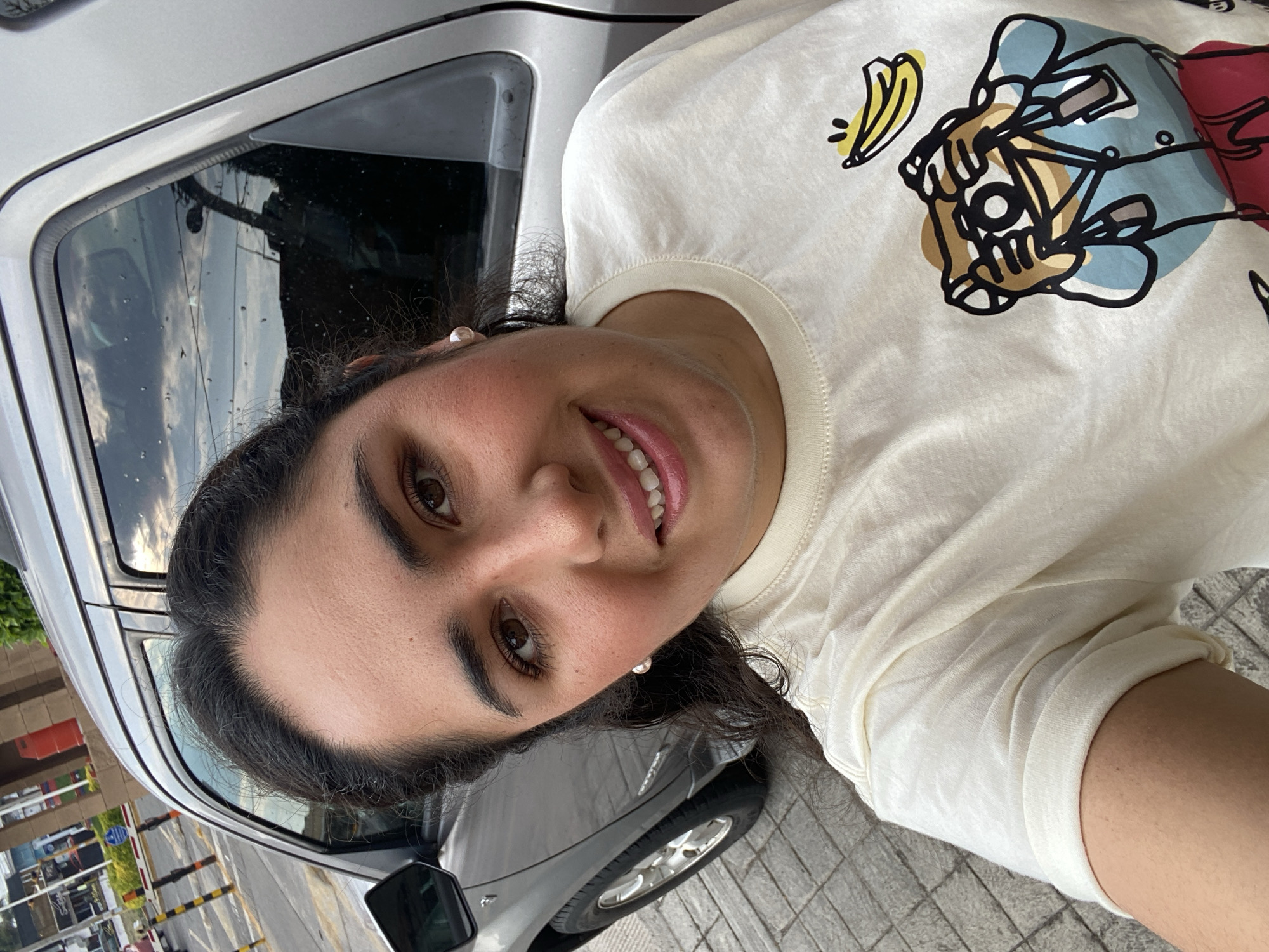 A woman with dark hair and brown eyes smiles towards the camera. She wears a light-colored t-shirt with a graphic design. Behind her is a silver car, and beyond that, a glimpse of a street with a barrier and some buildings. The location appears to be an urban environment, potentially San Bernardino Tlaxcalancingo, Mexico.