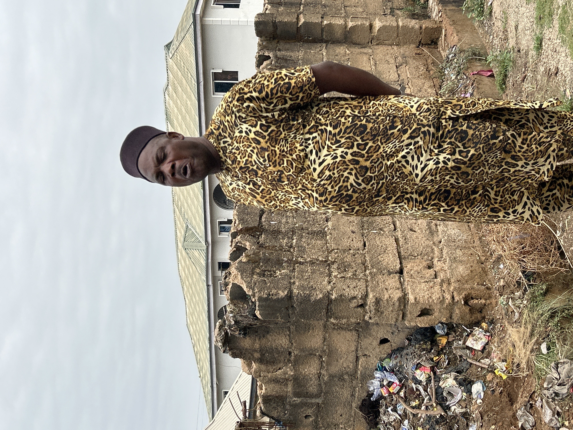 A man wearing a brown cap and a leopard-print shirt is standing in front of a dilapidated brick wall. The wall is partially crumbling and has debris and garbage at its base. In the background, there is a multi-story building with a silver roof. The sky is overcast. The location appears to be a neglected area in Jos, Nigeria.