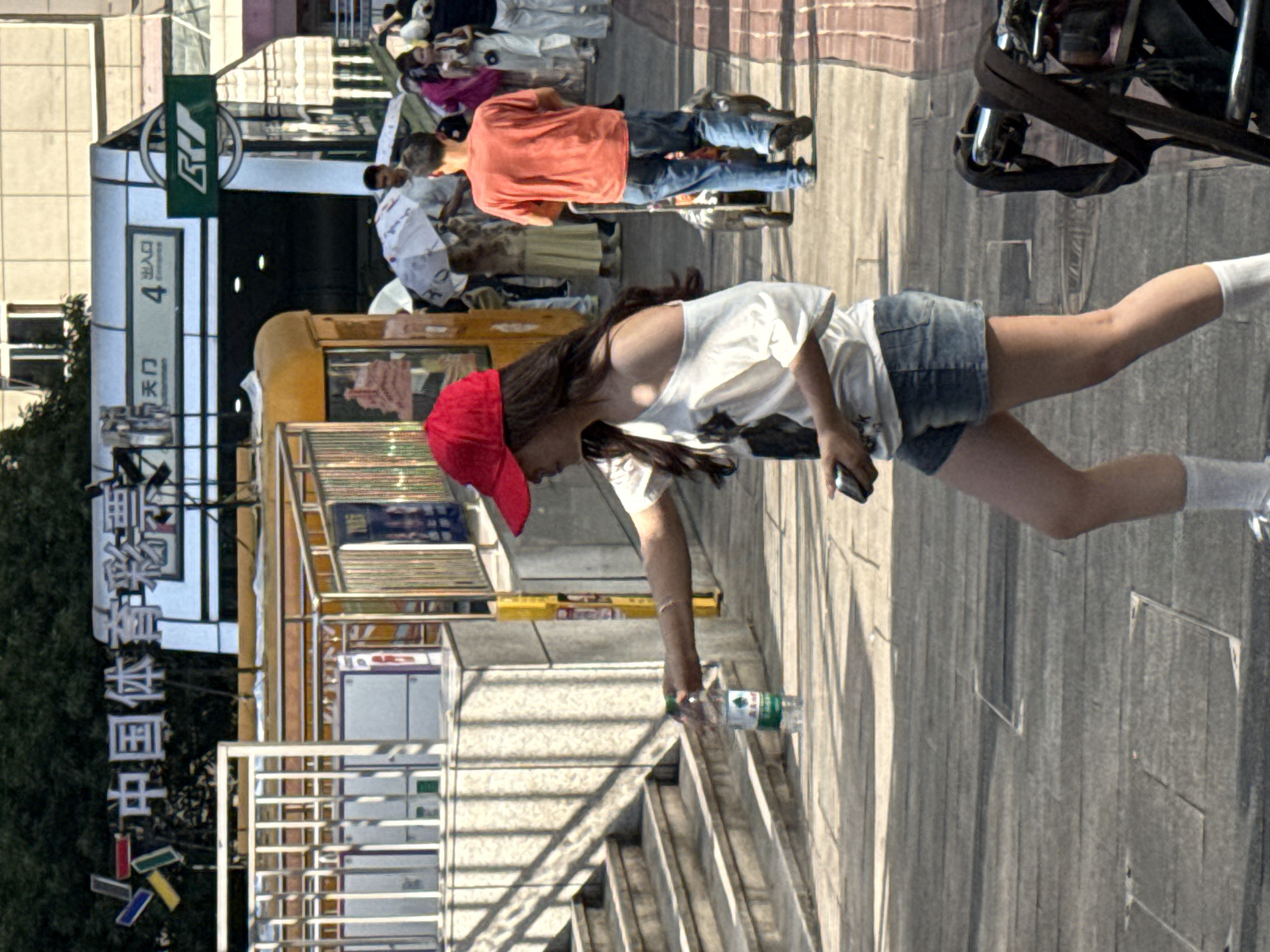 In Yuzhong District, China, a young woman wearing a red baseball cap, a white off-the-shoulder shirt, and denim shorts walks on a sidewalk. She is holding a plastic bottle of water in her right hand and a black mobile phone in her left. She is looking down and to her right. Behind her, several people are visible, some holding umbrellas. To the left of the woman, there are stone steps leading up to an entrance. In the background, there is a building with signs in Chinese. The overall scene depicts a busy urban environment.