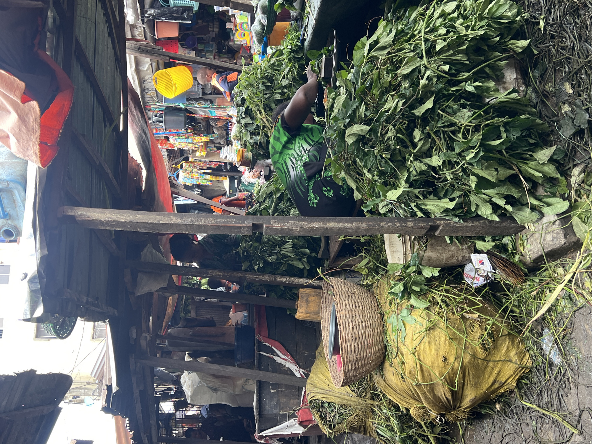 This is a medium shot of a market scene in Nkpor, Nigeria. The foreground is dominated by large piles of fresh green leafy vegetables, some of which are spilling out of large yellow sacks on the left. A woven basket containing a dark bowl is placed on top of one of the sacks. In the center-right of the image, a person wearing a green and black patterned shirt is actively sorting through the leafy greens. They are positioned behind a wooden counter or stall. The background reveals the bustling environment of the market, with more stalls, various colorful items hanging, buckets, and other people partially visible. A young child in an orange and blue shirt is visible in the upper right background. The overall environment suggests an outdoor market with temporary structures made of wood and corrugated metal, draped with tarps and fabrics for shade. The ground is wet and appears to be dirt or concrete, with debris and fallen leaves scattered around.