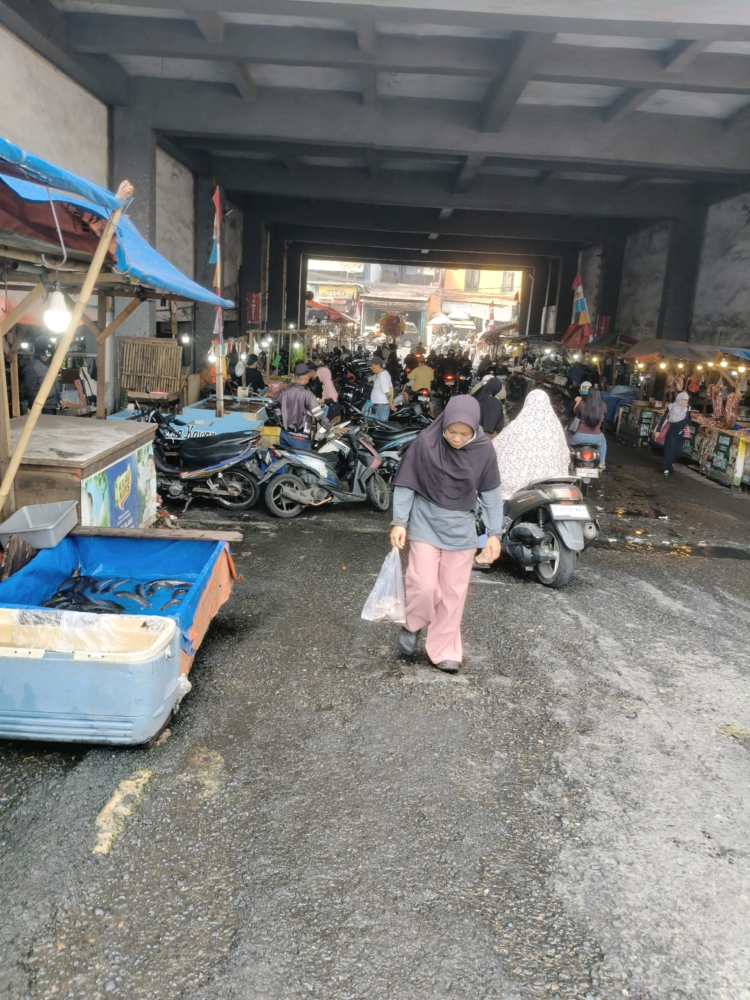 The image depicts an outdoor market beneath an overpass, likely a busy thoroughfare in South Tangerang, Indonesia, judging by the presence of motorcycles, stalls, and the general atmosphere. The foreground features a blue plastic tub filled with fresh fish, suggesting a fish market section. To the right of the tub, a woman wearing a hijab and pink trousers walks with a plastic bag in her hand, her gaze directed downwards. Behind her, several motorcycles are parked, and other market-goers can be seen amidst stalls displaying goods, some illuminated by hanging lights. The ground is wet and appears to be asphalt, indicative of recent rain or general market conditions. The overpass structure looms overhead, casting shadows and creating a confined yet open space for the market activities. The scene is bustling with commercial activity, with vendors and shoppers interacting within a vibrant urban environment.