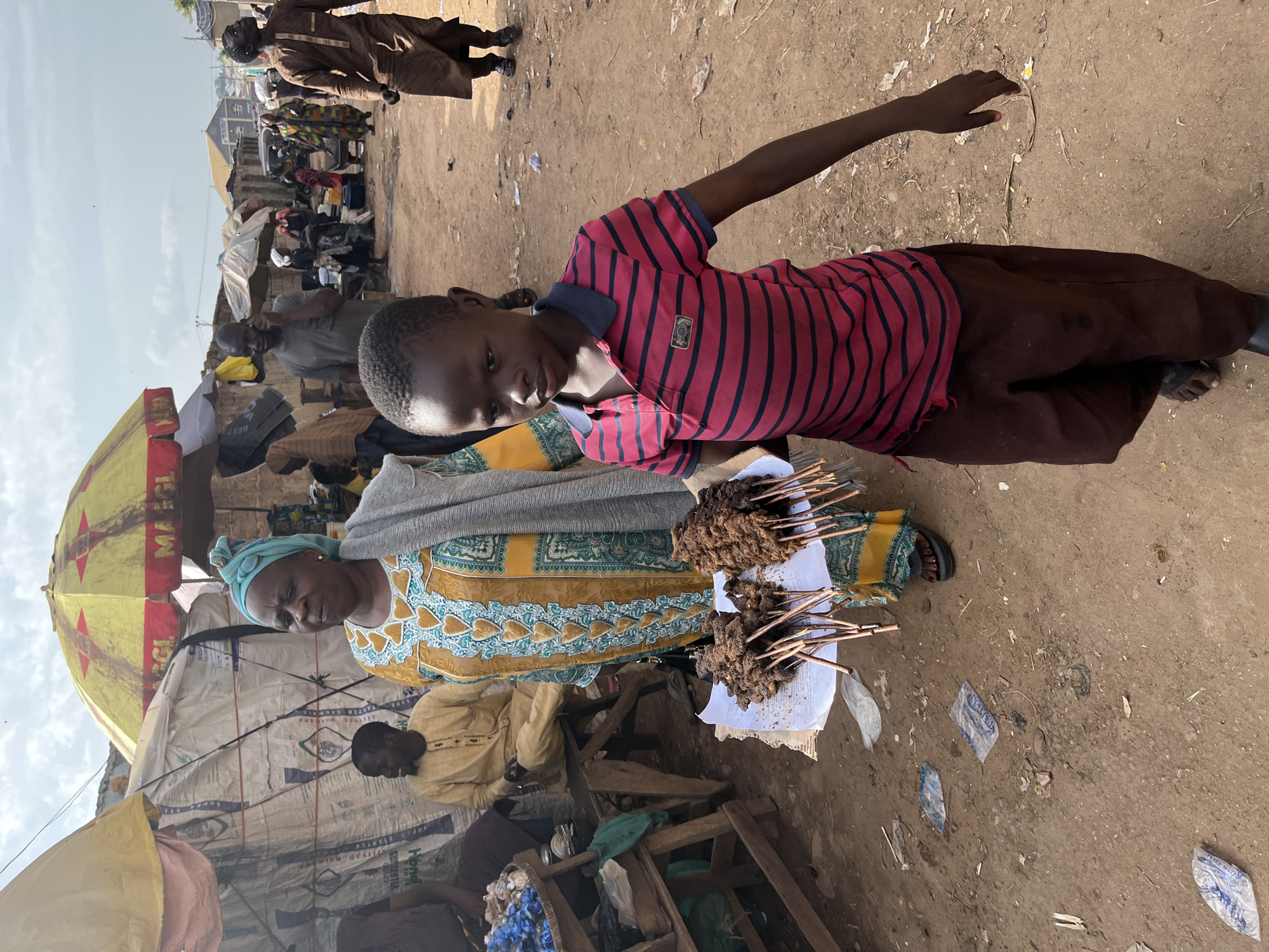 A street market scene in Rafin Par, Nigeria, features a young boy holding a tray of skewered meat in the foreground. He is wearing a red and black striped polo shirt and brown trousers. To his left, an older woman, dressed in a yellow and blue patterned robe and a turquoise headscarf, carries a gray shawl. Behind them, stalls are set up with various goods, including large sacks of what appear to be grains and a basket of blue items, possibly beads or fabric. Other people are visible in the background, some walking, others engaged in conversations, and a man in a brown garment is walking away from the camera. The ground is dirt, with scattered debris and plastic packaging. A yellow and red umbrella is prominent above some of the stalls. The sky is overcast.