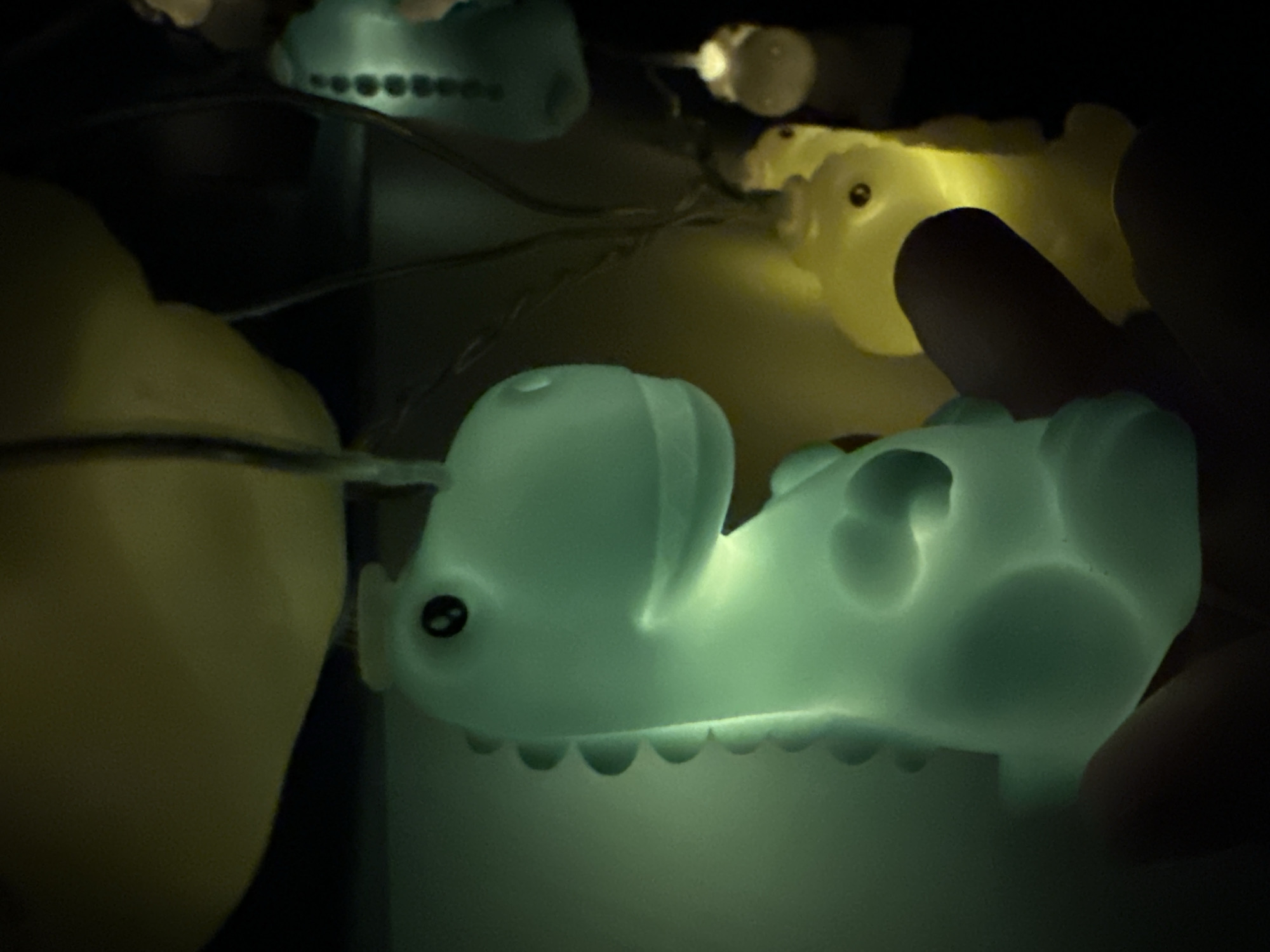 The image shows a string of glowing, stylized animal lights, possibly fairy lights, in a dimly lit environment. Predominantly visible are light-up figures resembling pigs or hippopotamuses, emitting a soft greenish-blue and yellow light. The lights are interconnected by a wire. Part of a hand intrudes on the right side of the frame. The location is unknown.