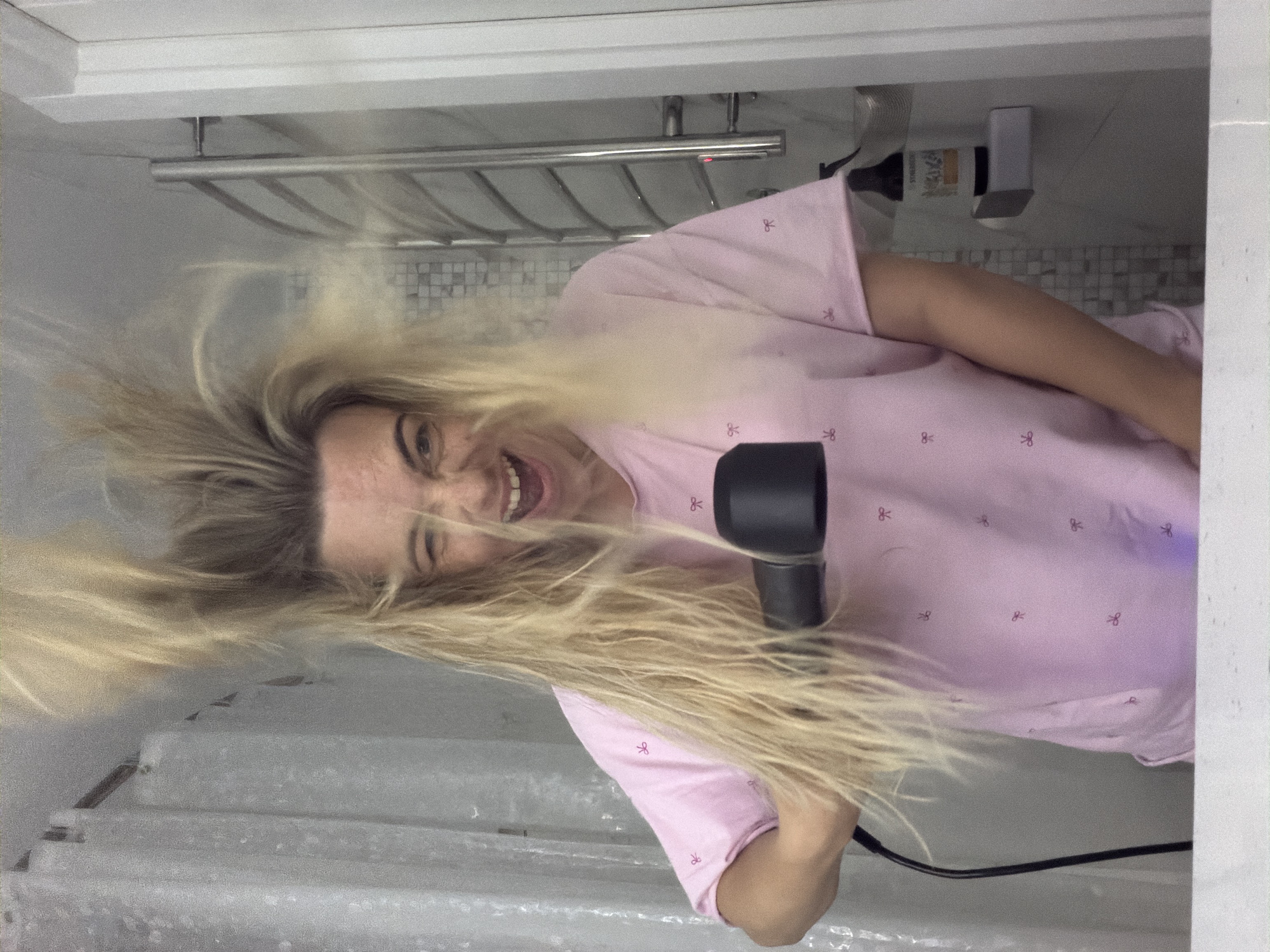 A woman with long blonde hair is using a black hairdryer. She is wearing a light pink shirt with bow designs. The setting appears to be a bathroom, as suggested by the bathtub and towel rack in the background. There is a bottle and a dispenser attached to the wall, also indicating a bathroom setting. The image provides no information to confirm its location in Votkinsk, Russia.