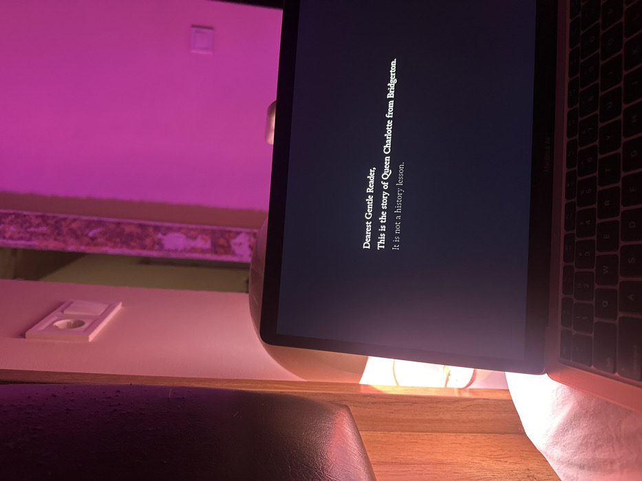 A laptop screen displays the opening text for "Queen Charlotte: A Bridgerton Story": "Dearest Gentle Reader, This is the story of Queen Charlotte from Bridgerton. It is not a history lesson." The laptop's keyboard is visible on the right. The laptop sits on a wooden surface, illuminated by a warm light. The background features a wall with pink lighting and decorative trim.