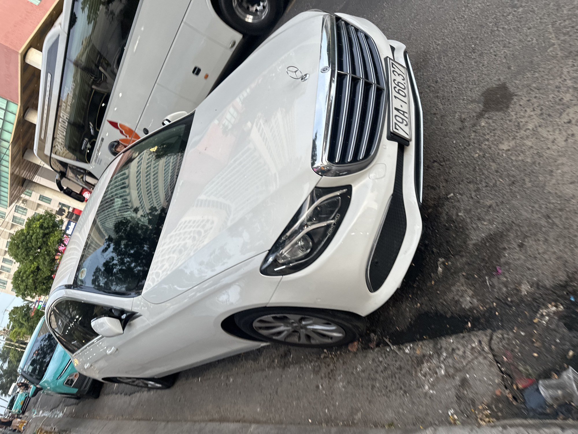 The image shows a white Mercedes-Benz E-Class sedan parked on the side of a street in Bac Nha Trang Ward, Vietnam. The car is facing forward, with its front bumper and grille prominently visible. The license plate reads "79A-166.37". Behind the Mercedes, a white Hyundai bus is also parked. Further down the street, a teal-colored car is visible. The street appears to be urban, with buildings and trees in the background. The ground is paved with asphalt and shows signs of wear and tear, with some scattered debris. The lighting suggests it is daytime, possibly late afternoon.