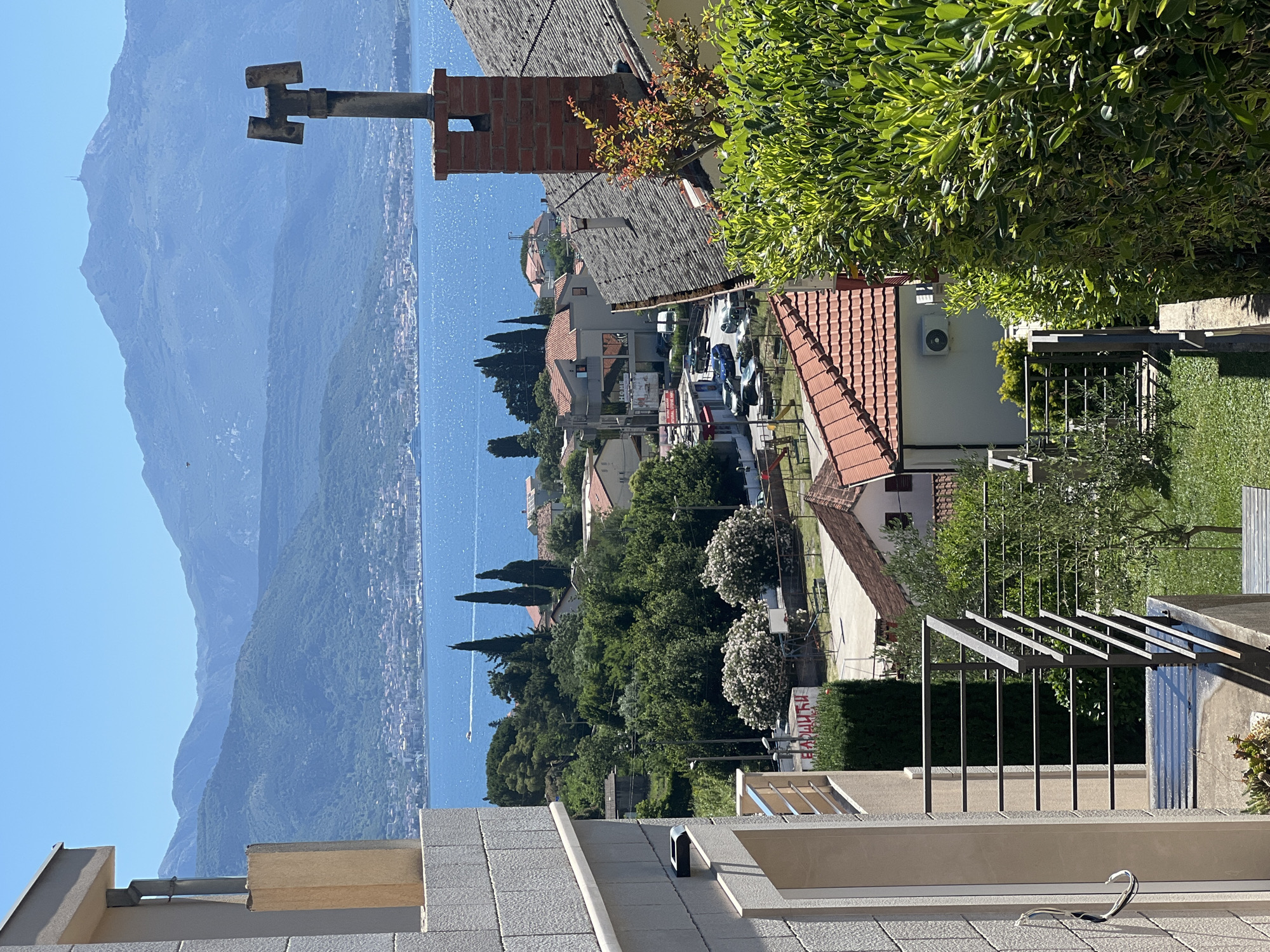 The image shows a view overlooking the city of Baošići, Montenegro. The scene is dominated by a body of water, likely the Bay of Kotor, with mountains visible in the background. Buildings with red-tiled roofs are interspersed with trees. A structure with a metal railing is in the foreground. A chimney with a weather vane is visible to the left.